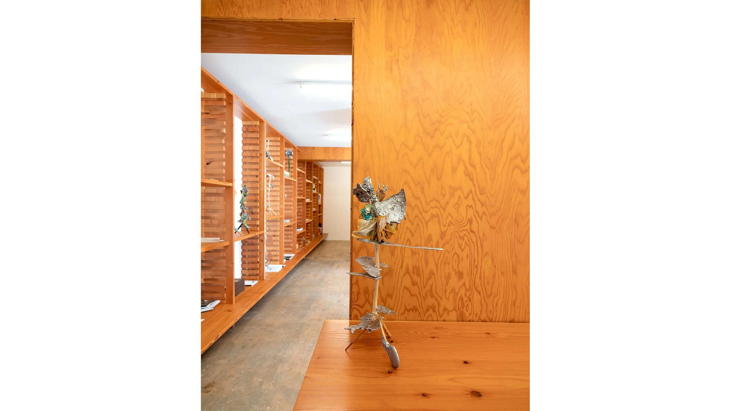 Yasue Maetake Three-Legged Idols gallery installation: single three-legged sculpture on wooden surface in foreground with long wooden bookshelf corridor visible in the background
