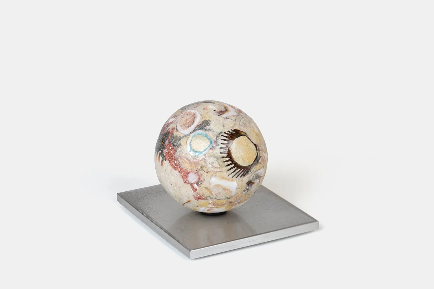 Yasue Maetake Orb Materia—Bird Tracks, 2026. Spherical form with soft beige mineral surface containing circular shell fragments and a dark radial impression, resting on a square aluminum base.