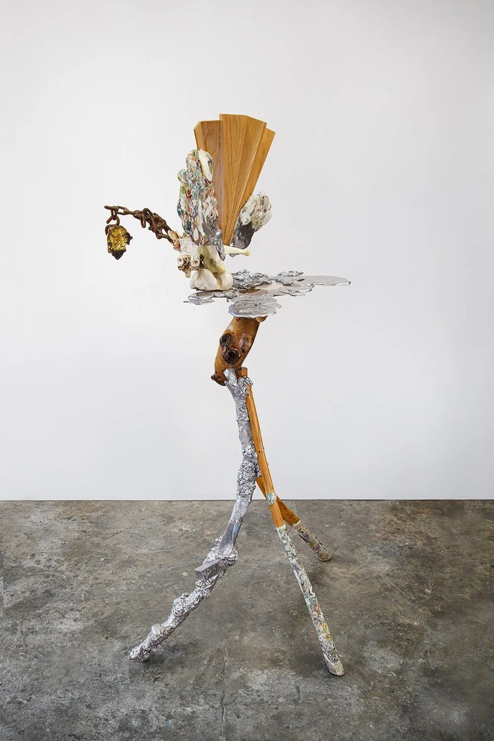 Yasue Maetake, Primordial Soup, 2023-2025. Tall hybrid sculpture on a three-legged tripod base with a central wooden support, light wooden fan structure, and upper assemblage of textured organic and metal forms with a hanging branch element.
