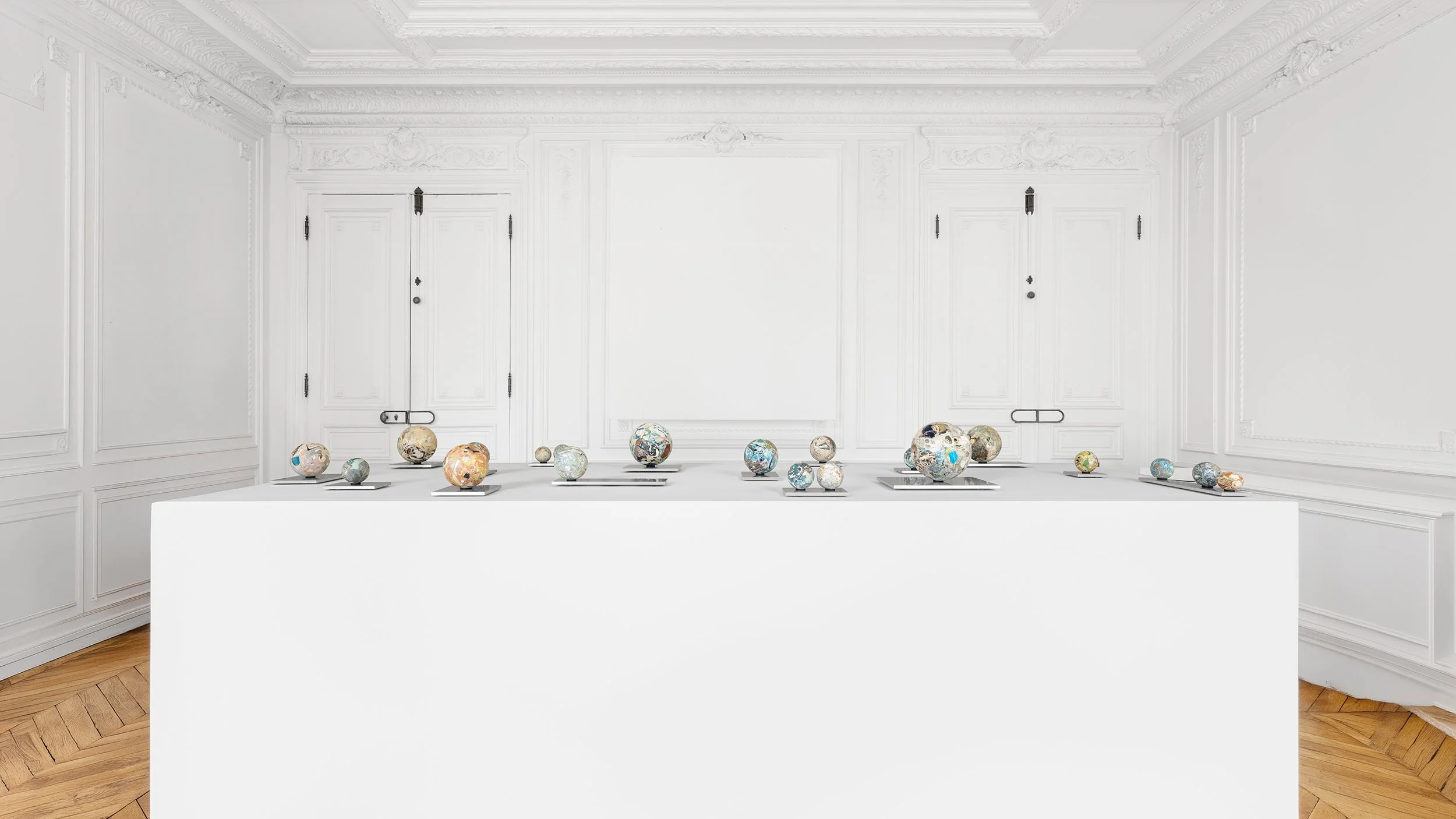 Yasue Maetake Orb Materia—Carved Algos gallery installation: wide frontal view of multicolored carved spheres arranged across a long white platform in a European classical white room