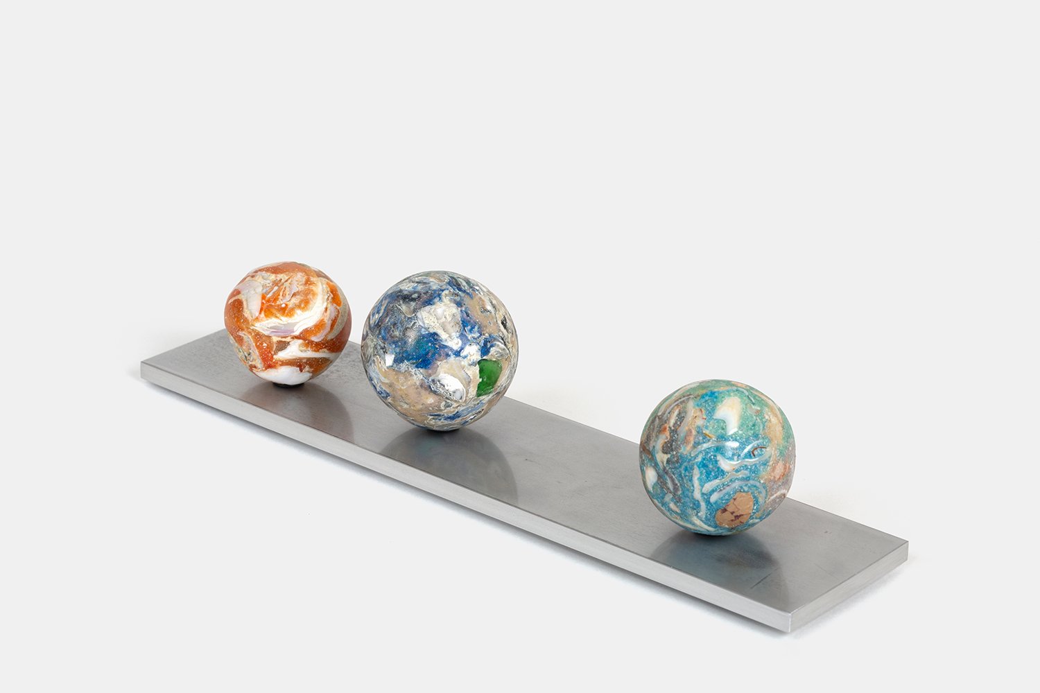 Yasue Maetake Orb Materia—Tangerine PCB, 2026. Three spherical sculptures arranged on a long rectangular aluminum base, showing orange, blue-white, and turquoise surfaces with embedded mineral and shell fragments from a side angle.