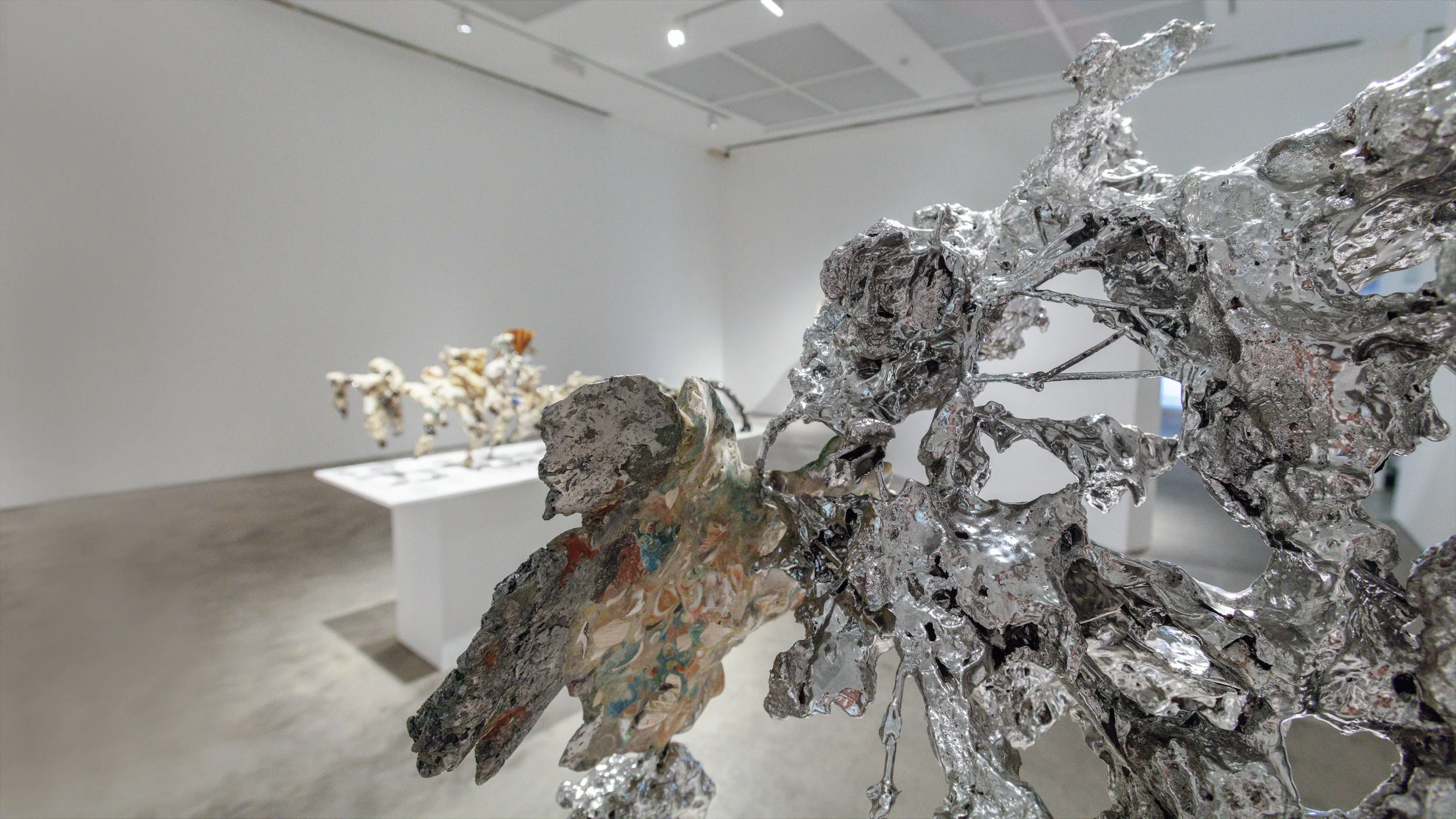 Yasue Maetake Mare Fecunditatis gallery installation: close foreground of complex silvery molten form with colorful accretions, another sculpture on white pedestal in the distant white gallery space