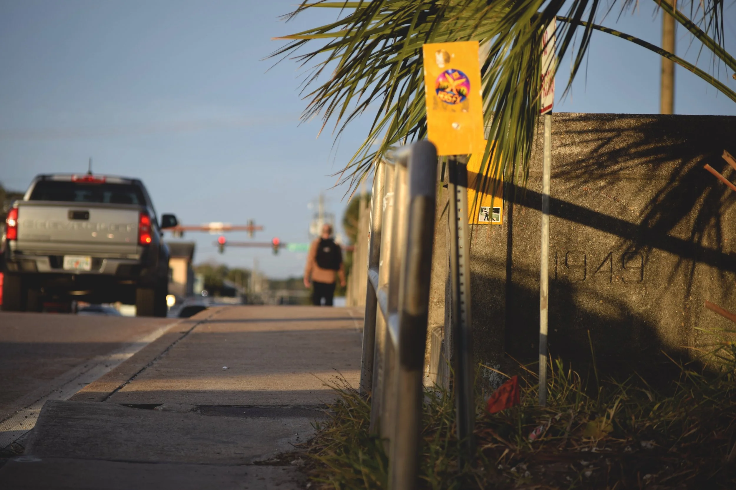 King Street Bridge in St. Augustine, Florida. A sidewalk next to a street with a metal guardrail, a yellow sign, and a palm tree shadow on a concrete wall. In the background, a stop gate and a person walking, with a pickup truck driving by.