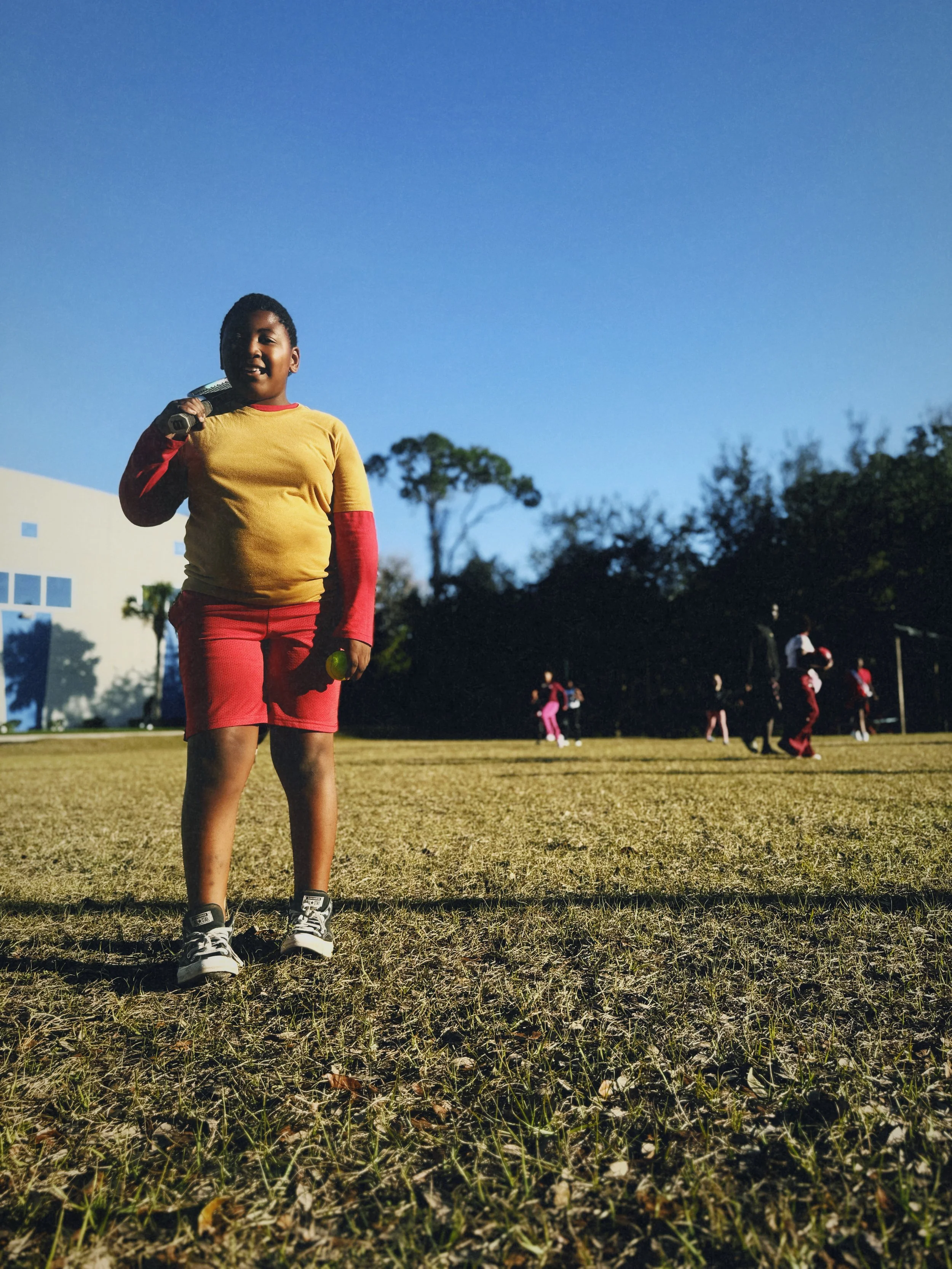 A young boy standing outdoors on a grassy field at the Boys & Girls Club in St. Augustine, Florida holding a tennis ball in one hand and a racket over his shoulder, with other children playing in the background.