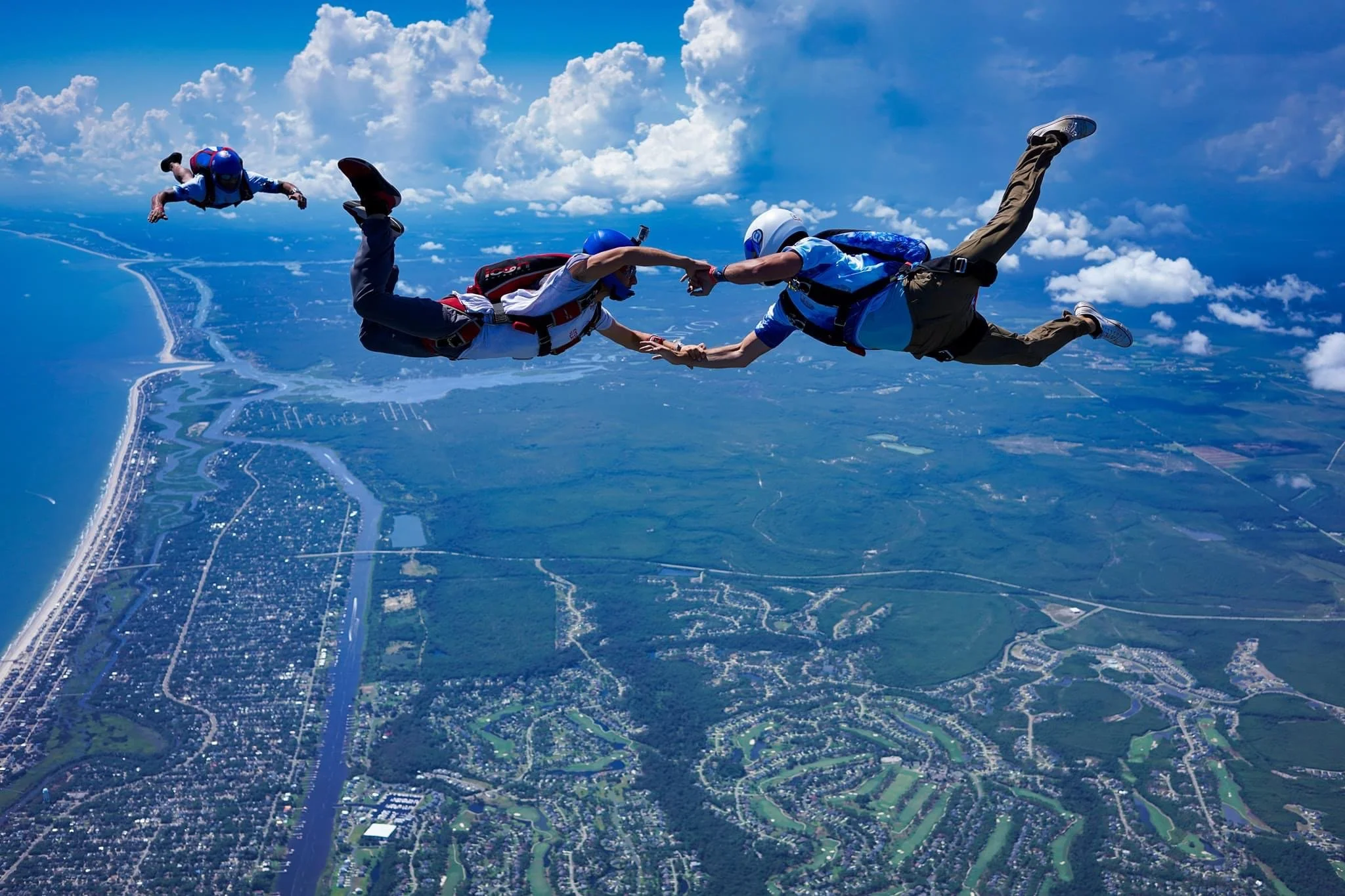 Two skydivers holding hands mid-air during a freefall over St. Augustine ih a beach, water, and green landscape, with another skydiver in the background. 