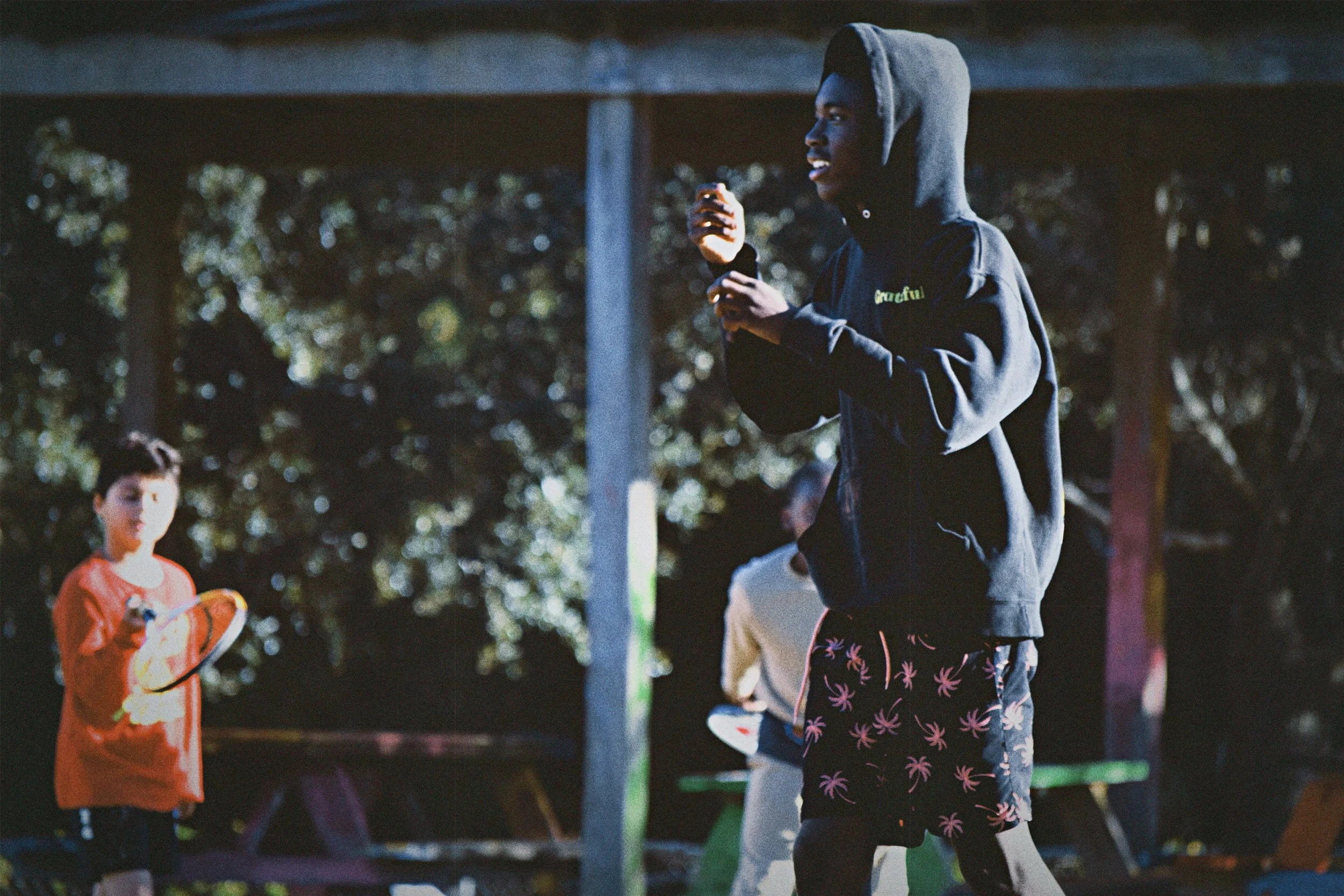 A group of children playing outdoors at Boys & Girls Club in St. Augustine, Florida, with one boy in the foreground holding a ball, wearing a dark hoodie and patterned shorts, while two other children are in the background.