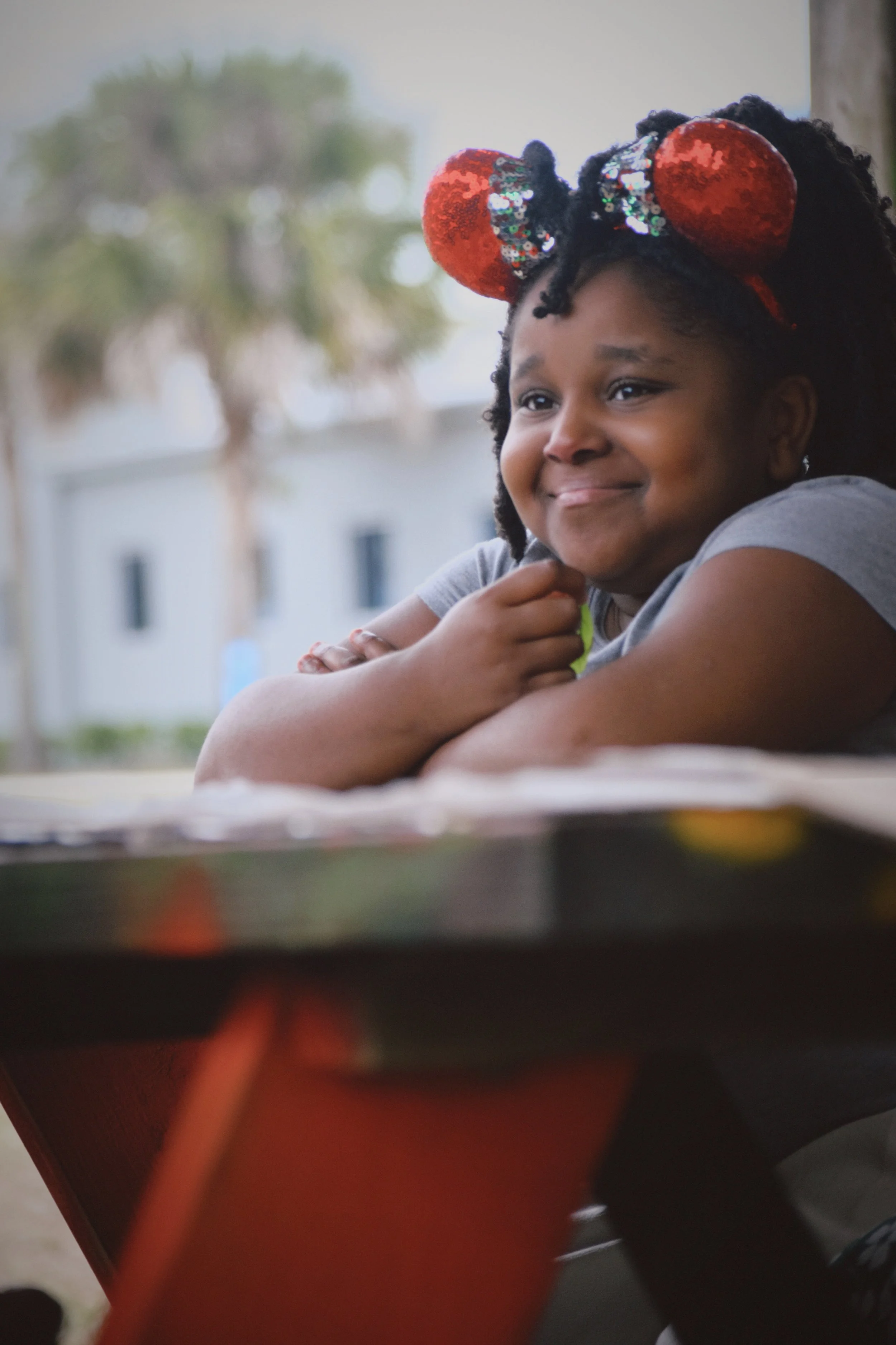 A young girl with curly hair and red sequined Minnie Mouse ears headband smiling and resting her arms on a table at Boys & Girls Club in St. Augustine, Florida, outside on a cloudy day with a blurred background of trees and buildings.
