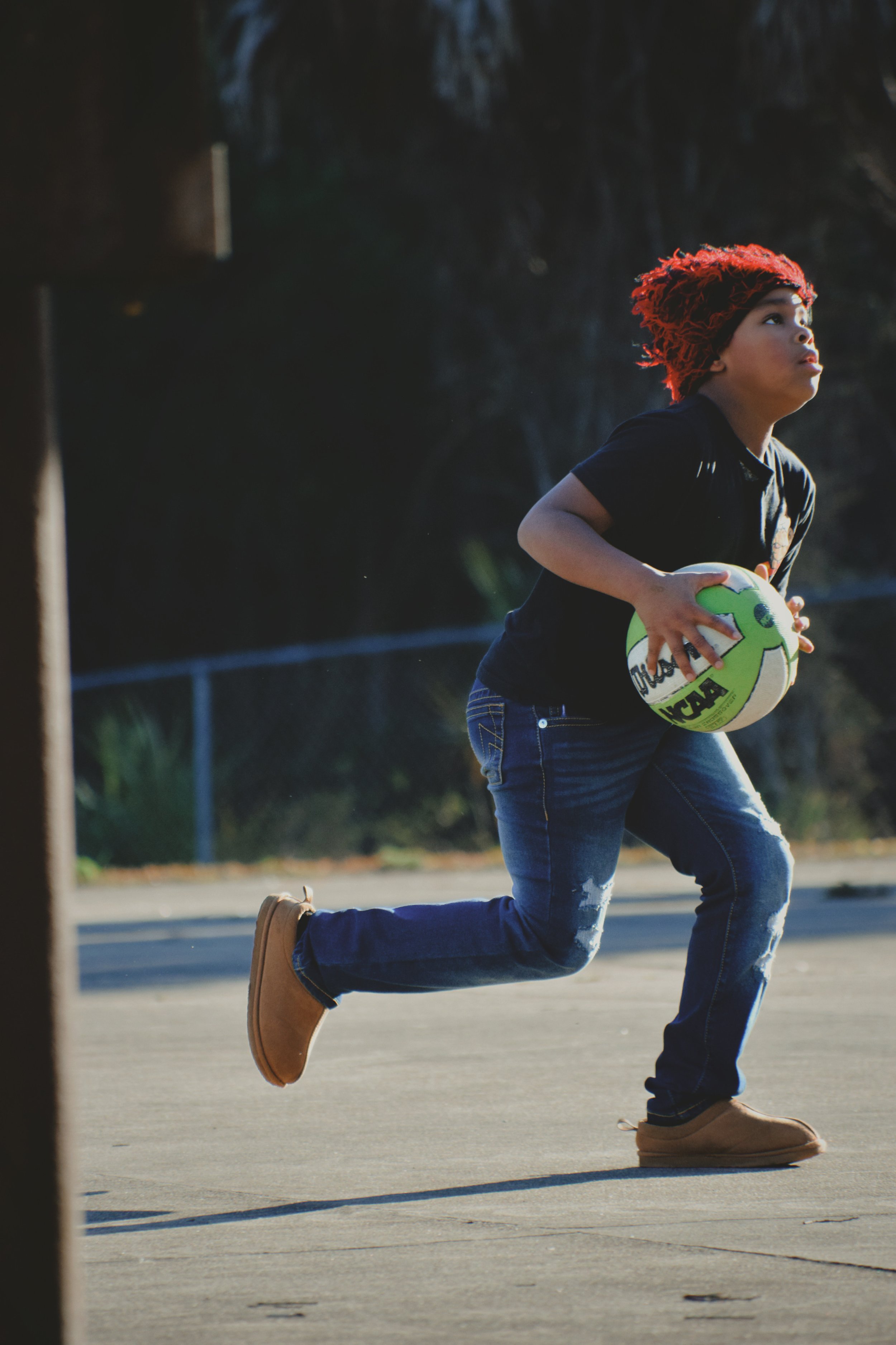 A young child running on basketball court at Boys & Girls Club in St. Augustine, Florida, holding a green and white basketball, wearing a red beanie, black shirt, blue jeans, and tan boots.