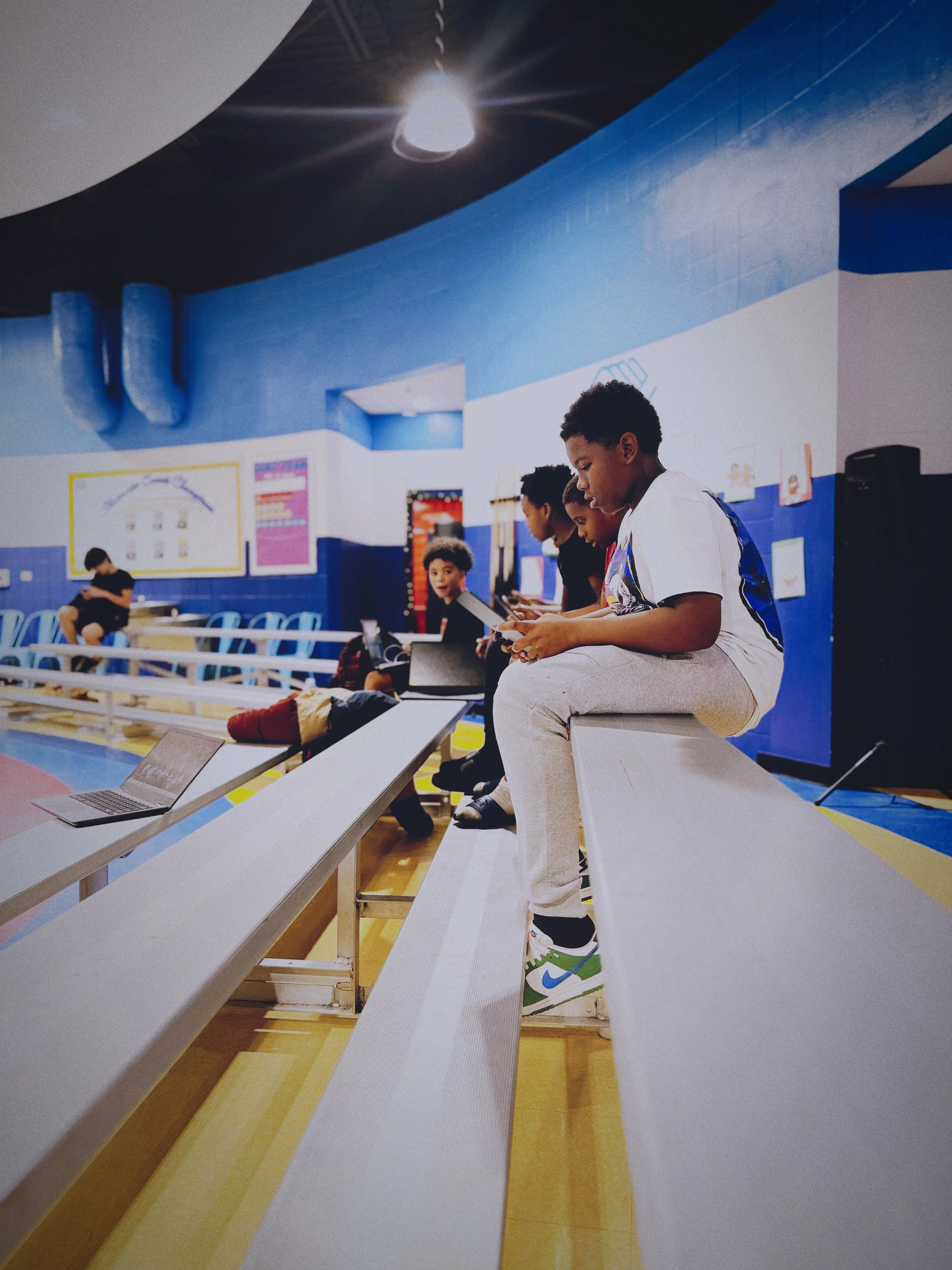 Kids sitting on bleachers in a gym at Boys & Girls Club in St. Augustine, using laptops and tablets, with a blue and white wall and colorful posters in the background.