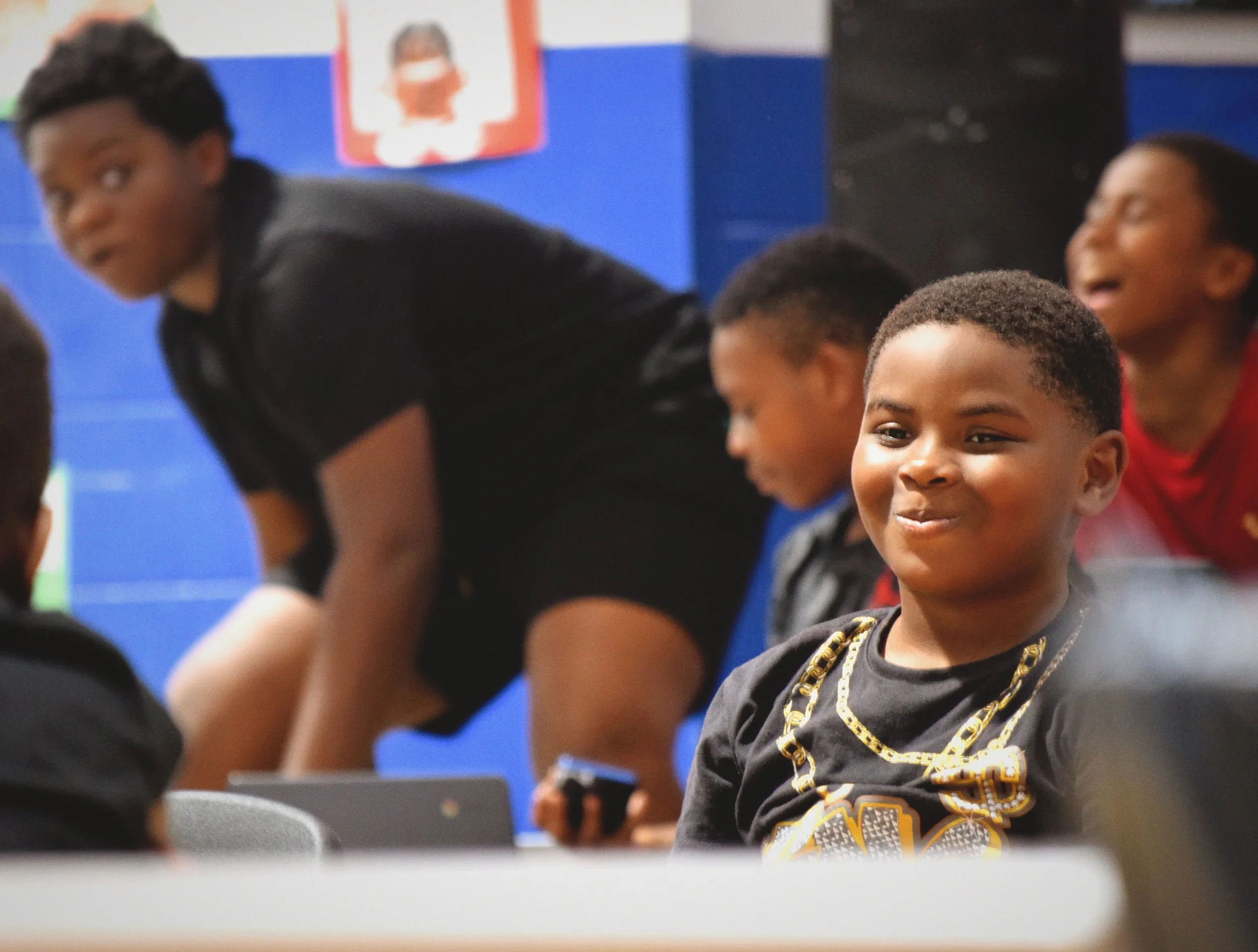 Young boy smiling, wearing a black shirt with gold chain design, at Boys & Girls Club in St. Augustine, Florida with other children and a woman leaning over, background features a blue wall and framed pictures.