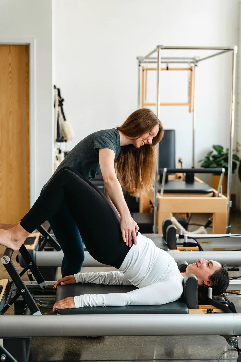 A woman assisting another woman during a Pilates exercise on a reformer machine in a studio.