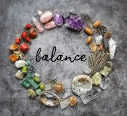 Healing Journey - Re-energizing and Self Awareness image showing a circle of colorful crystals, stones, feathers, and natural objects arranged around the word “balance” on a textured gray background.