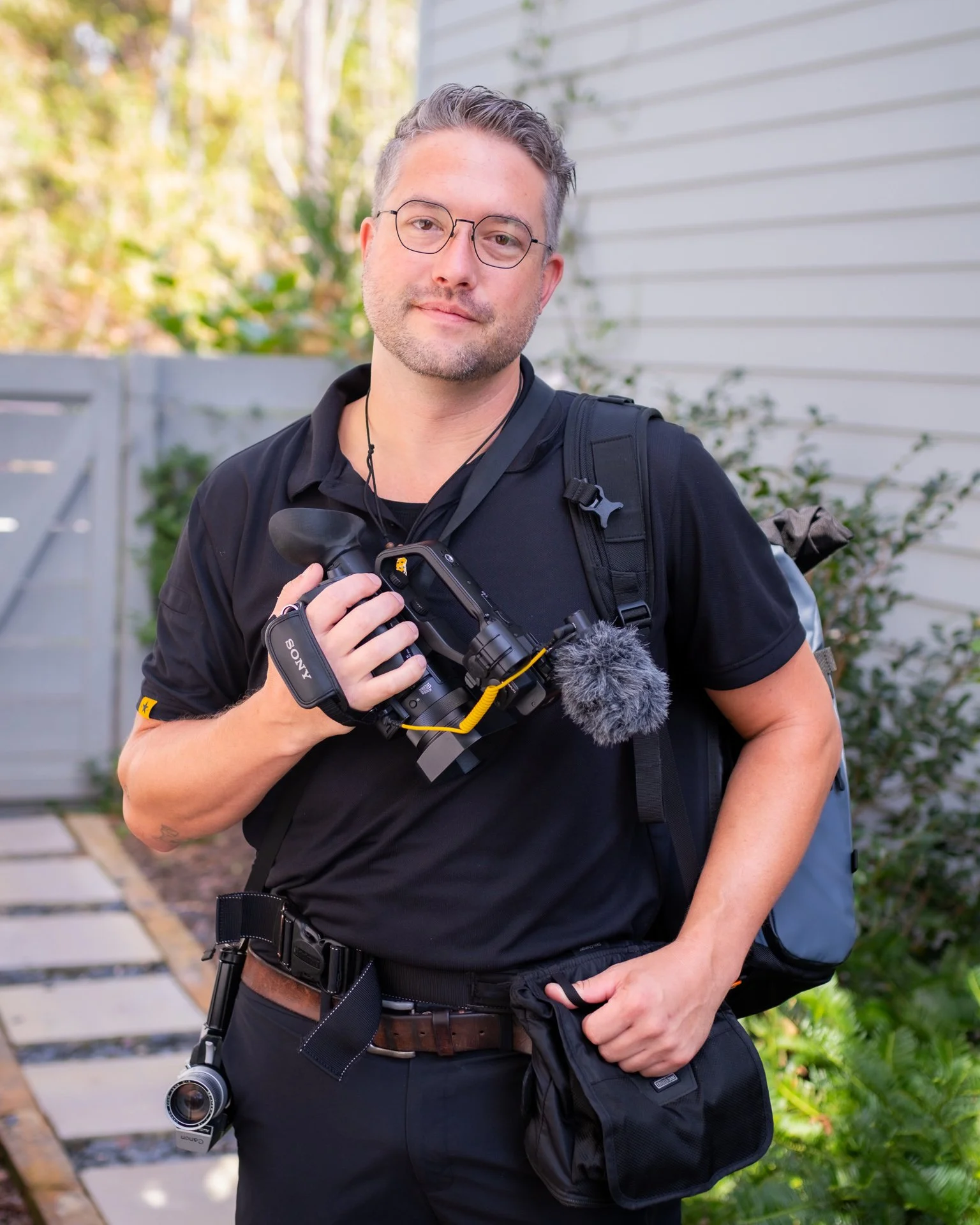 A man in black clothing holding a professional camera with a mounted microphone outdoors, standing next to a white fence and green shrubbery.
