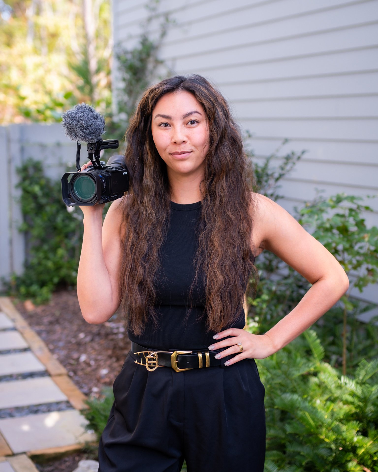 A woman with long, wavy brown hair holding a video camera with a microphone outdoors, standing on a garden pathway beside a gray house with greenery.
