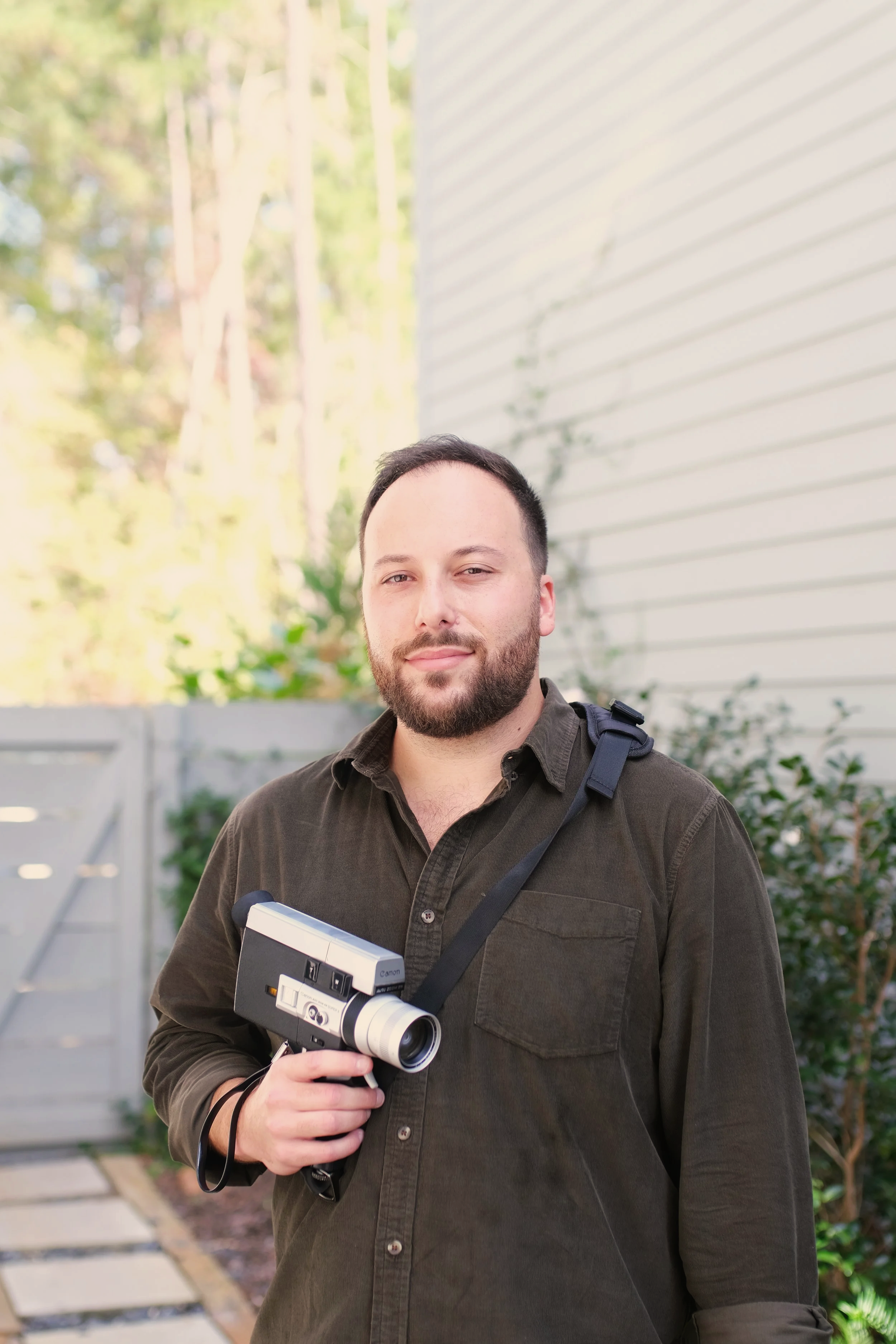A man with a beard holding a vintage film camera, posing for a photography with greenery in the background. Founder of For Keeps Film Co.