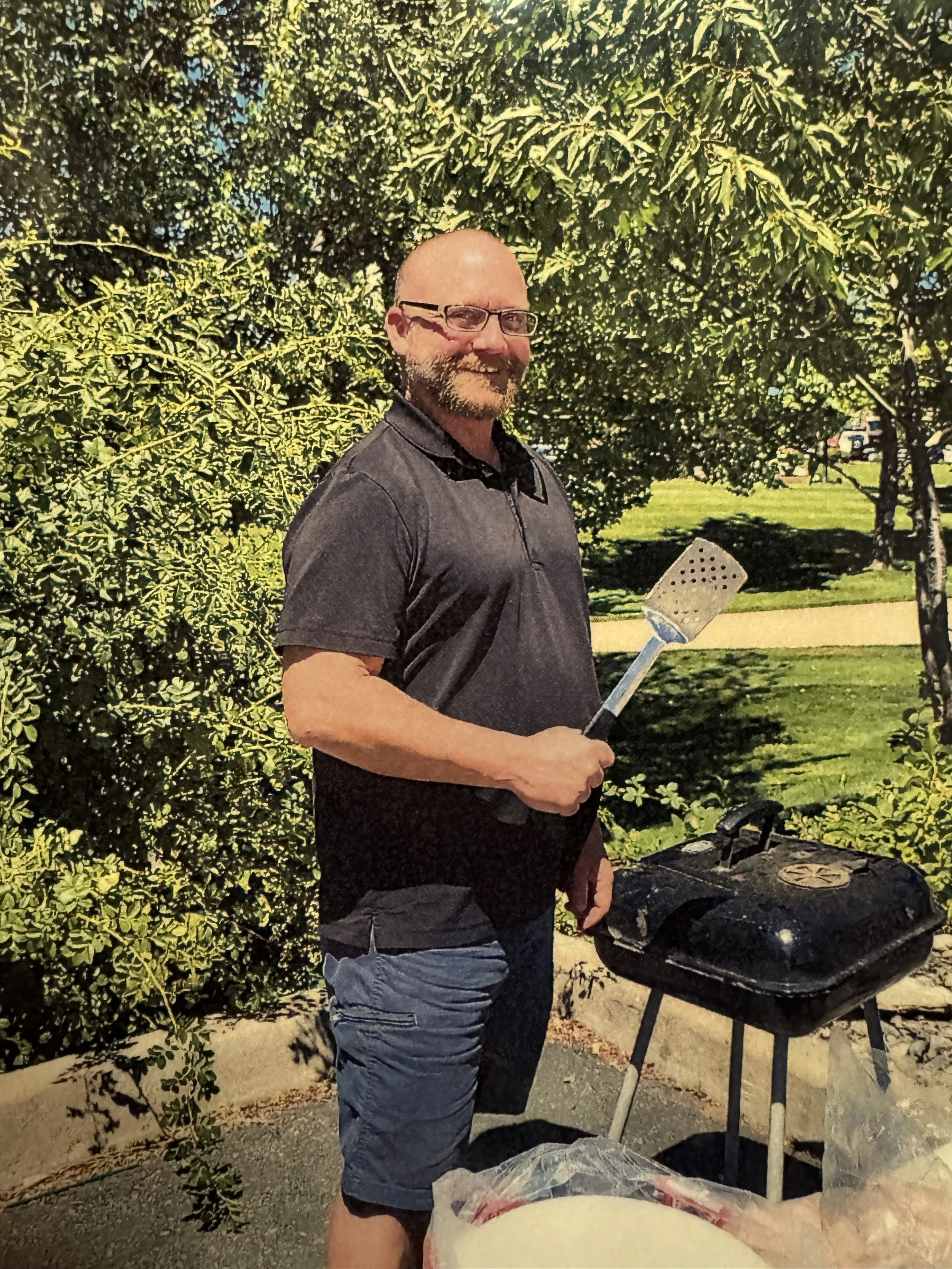 A man with glasses, a beard, and wearing a dark gray polo shirt and shorts, is standing outdoors near a small black grill with a lid. He is holding a spatula and is smiling. Behind him are trees and a green park.
