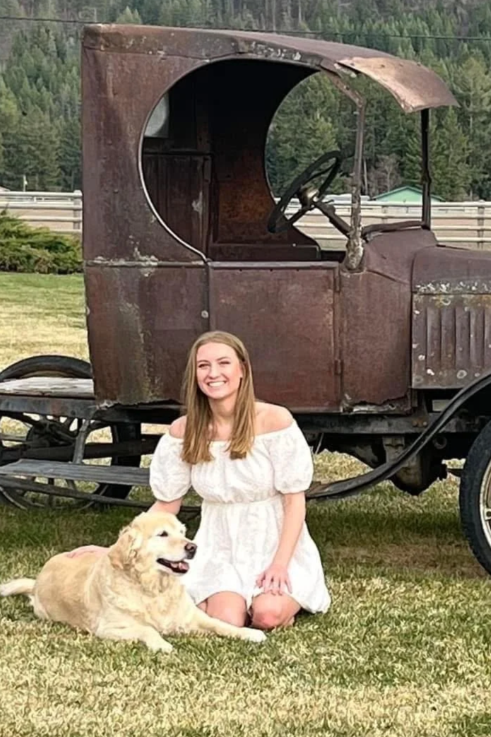 A young woman with long hair, wearing a white off-shoulder dress, kneeling on the grass next to a yellow dog lying down. They are in front of an old, rusty, vintage car with a covered driver's seat and no windows, parked on a grassy area with trees and a fence in the background.