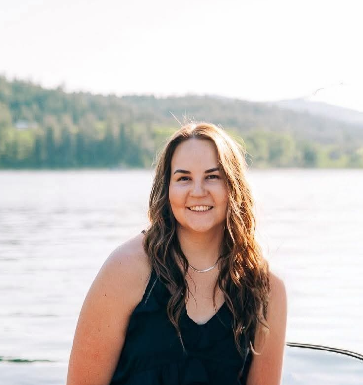 Young woman with long brown hair smiling near a lake with green hills in the background.