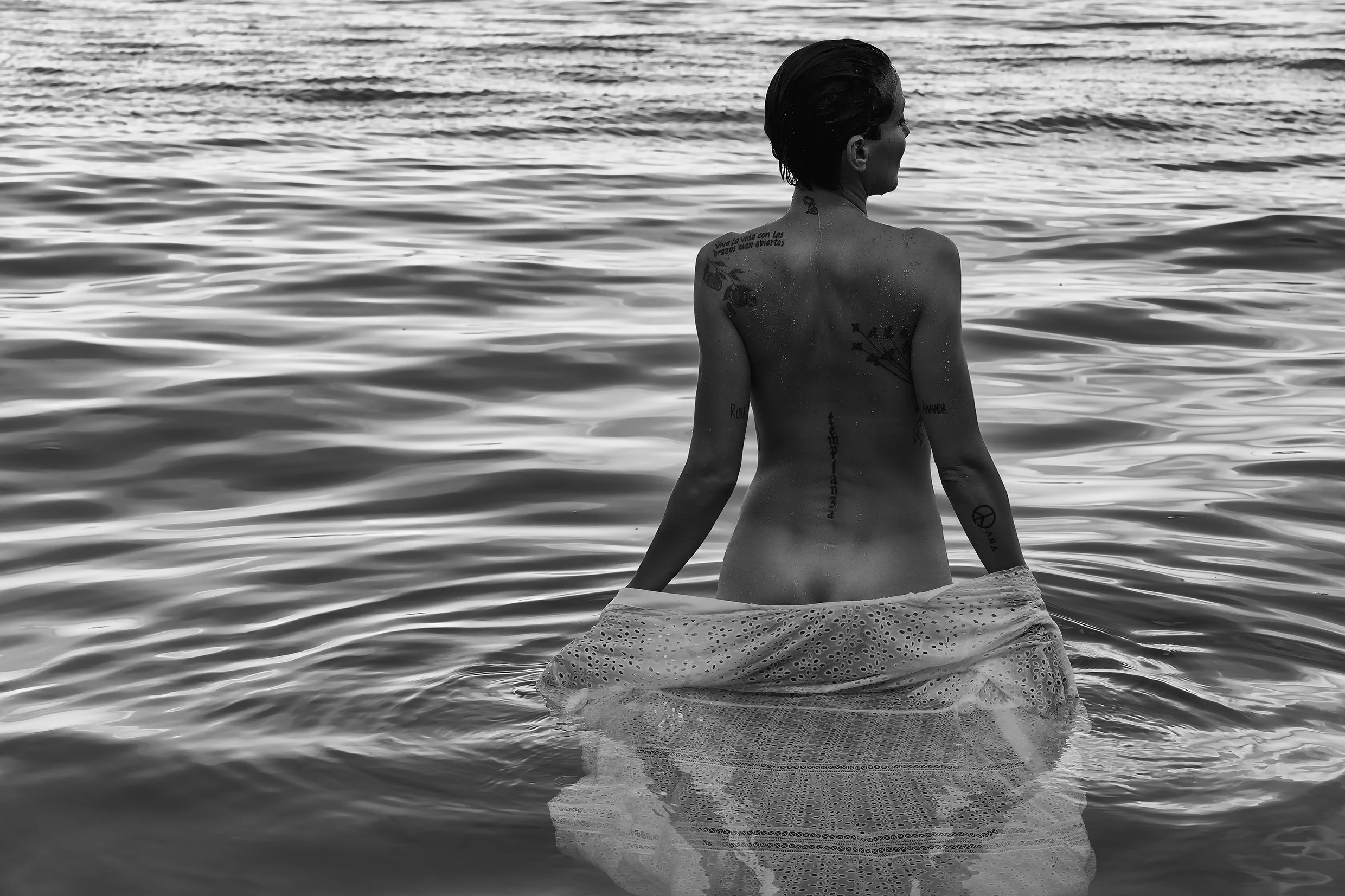 A woman with tattoos sitting in water at the beach, facing away from the camera.