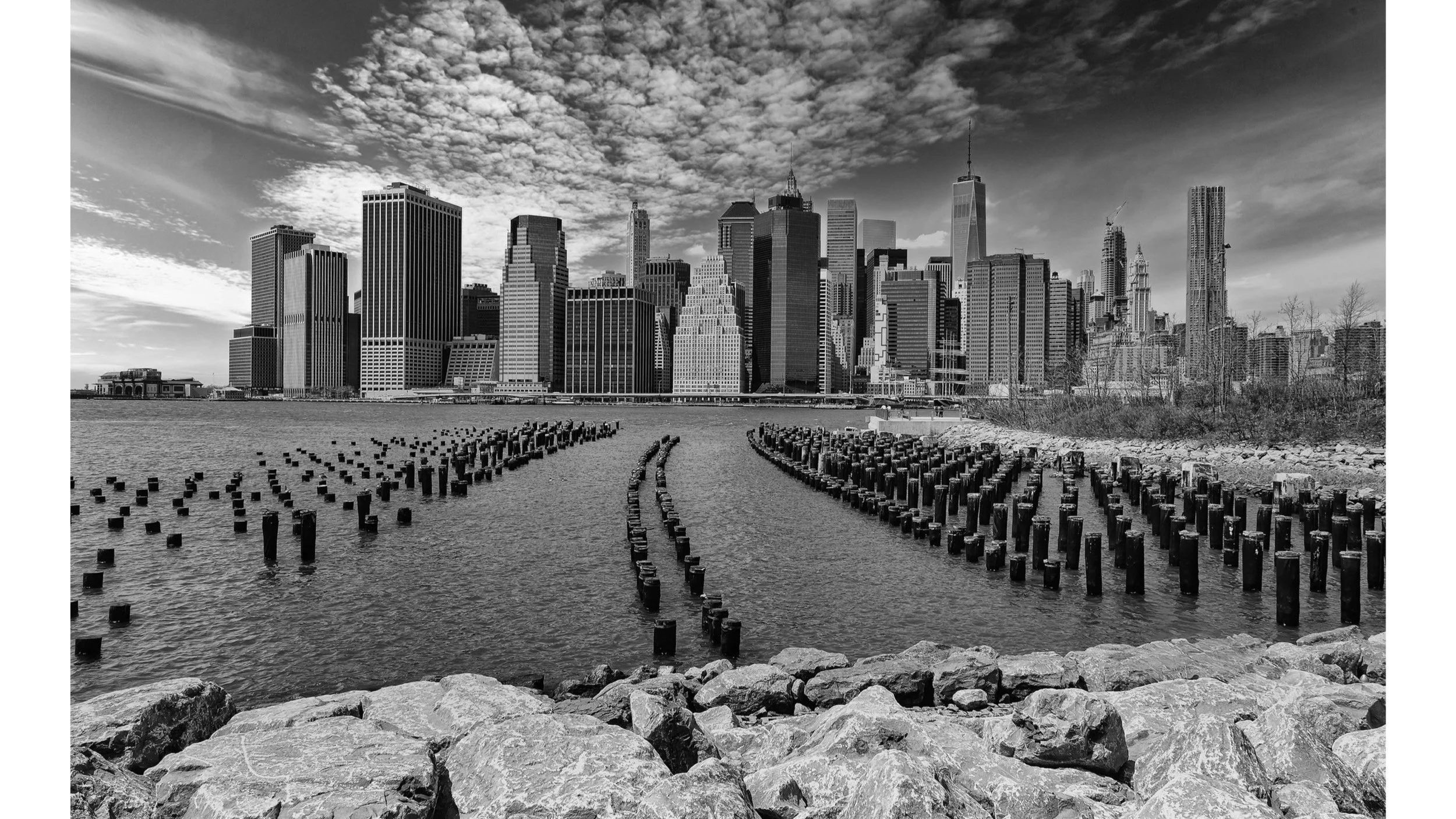 Black and white photo of the Manhattan skyline with skyscrapers, cloudy sky, and water in the foreground, featuring wooden pilings along the pier.