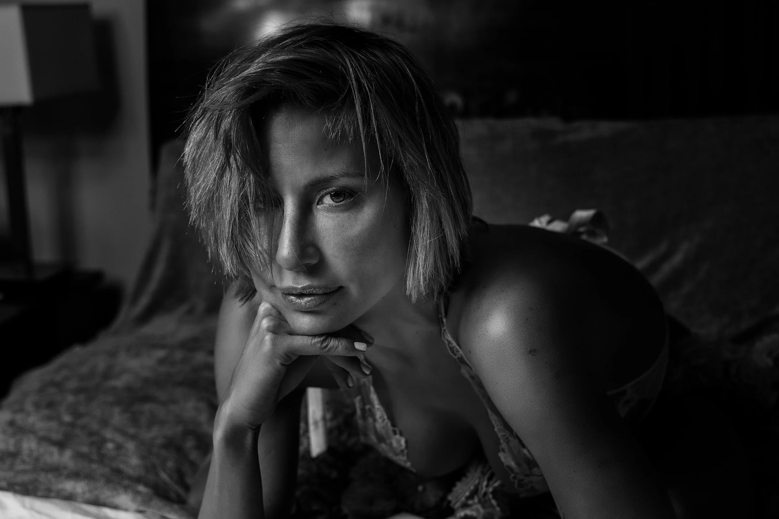 A woman with short, tousled hair lying on a bed, resting her chin on her hand, looking at the camera in a black-and-white photograph.