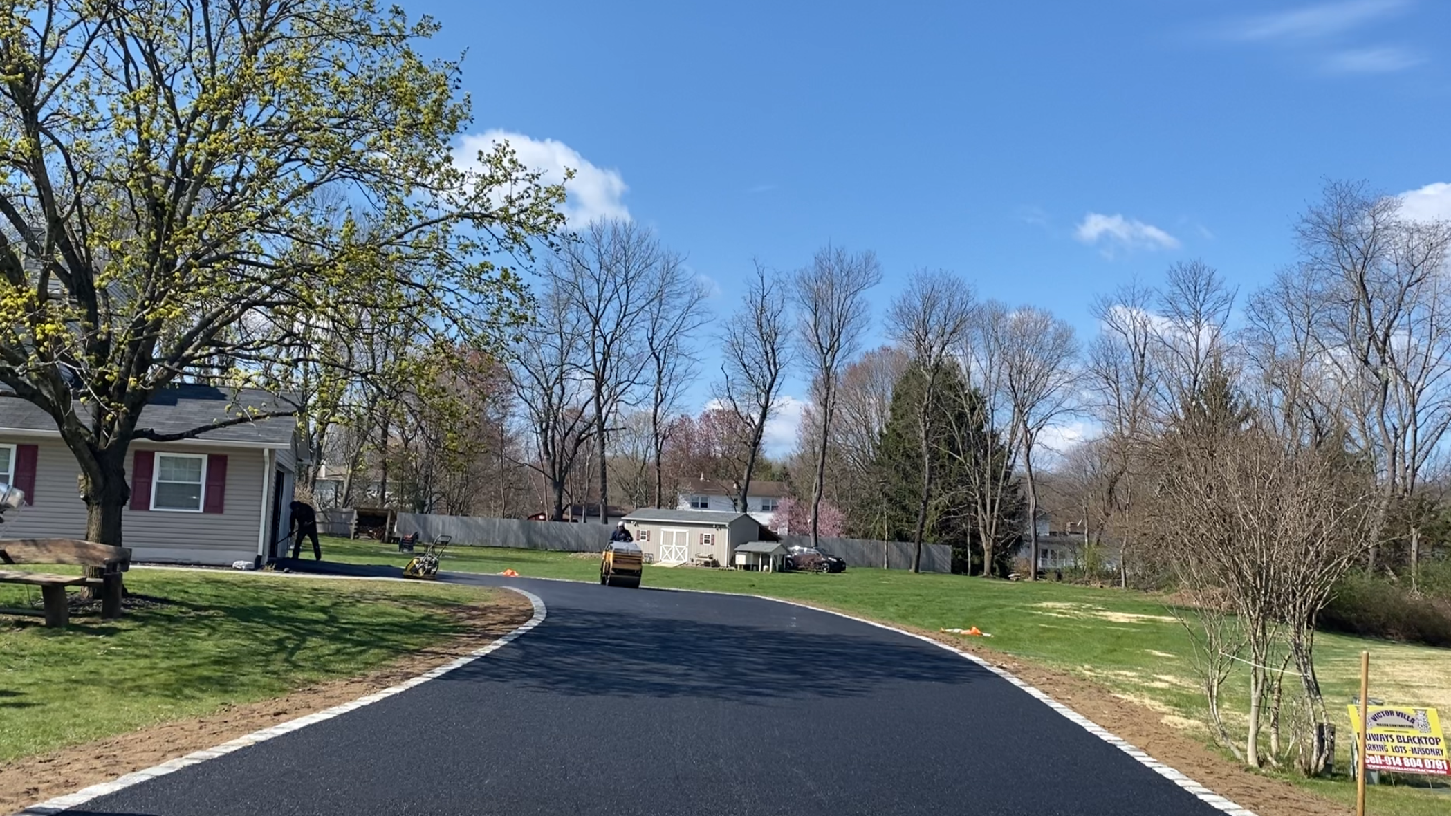 A newly paved black asphalt driveway curves through a residential yard on a clear day. There are trees with budding leaves, a small house with red shutters on the left, and a yard worker with equipment on the left. A shed and parked cars are visible