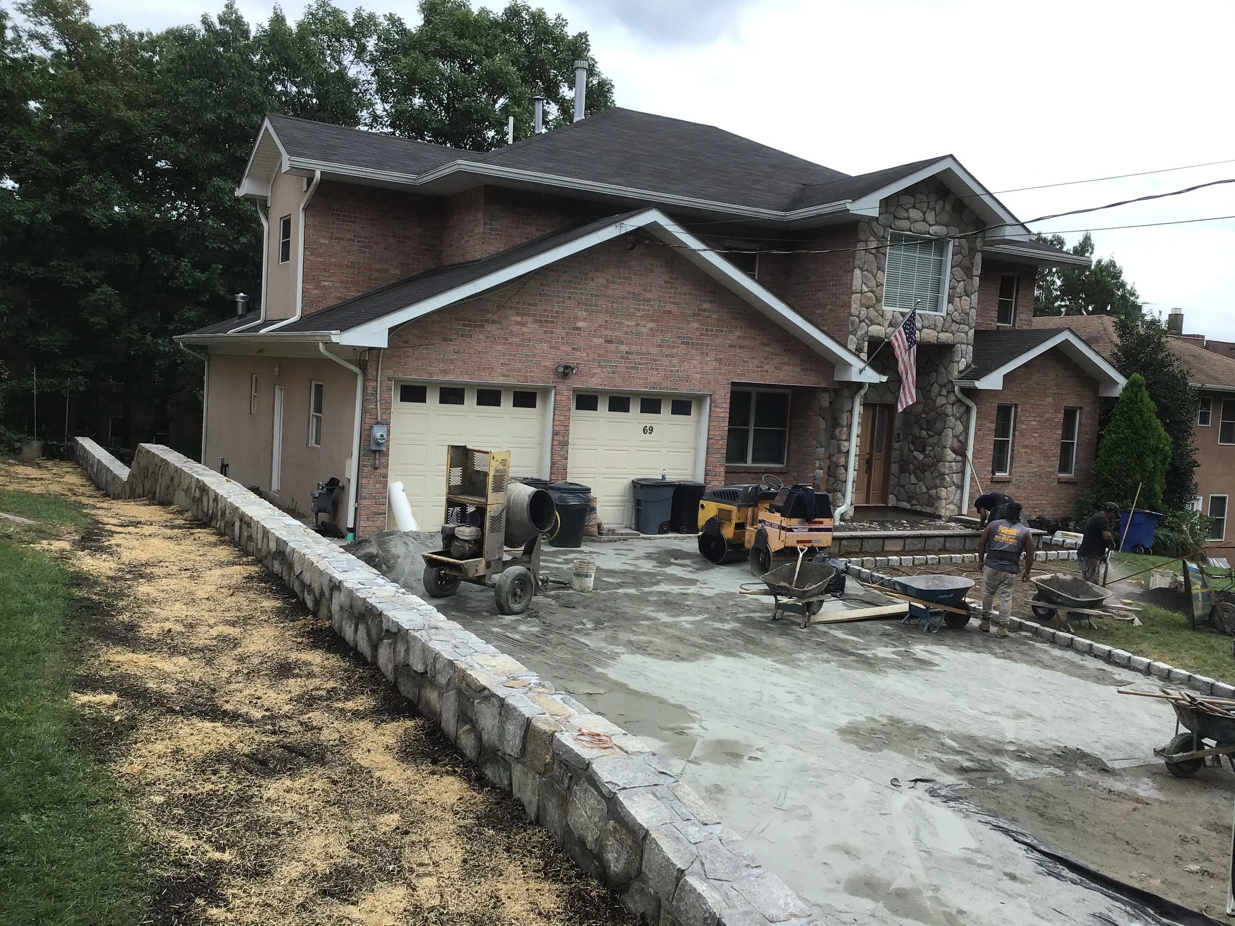 House under renovation with construction workers leveling a concrete driveway, with wheelbarrows, construction equipment, and a stone wall border.