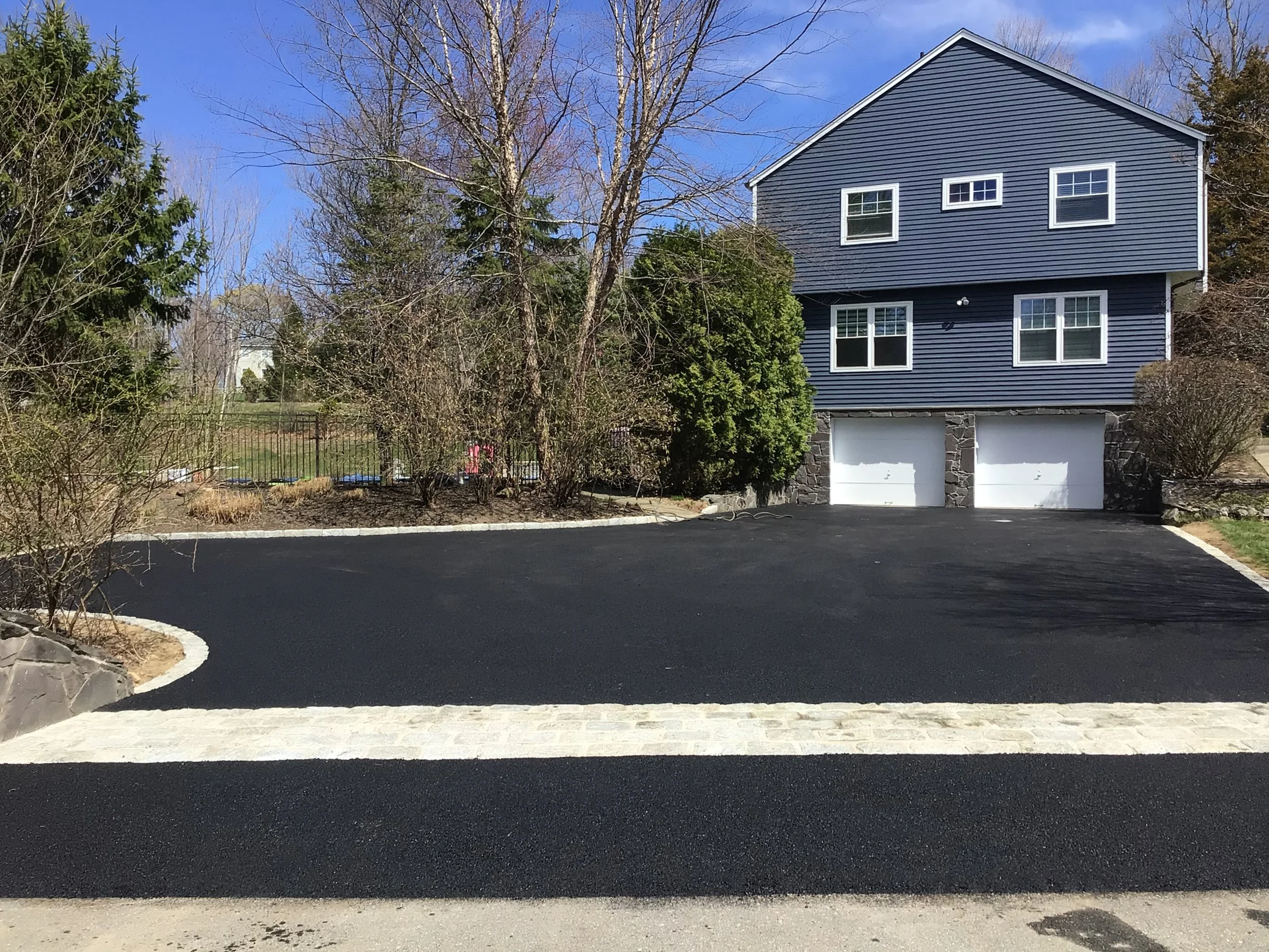 Newly paved black asphalt driveway leading to a house with dark blue siding and two white garage doors, with trees and bushes surrounding the property.