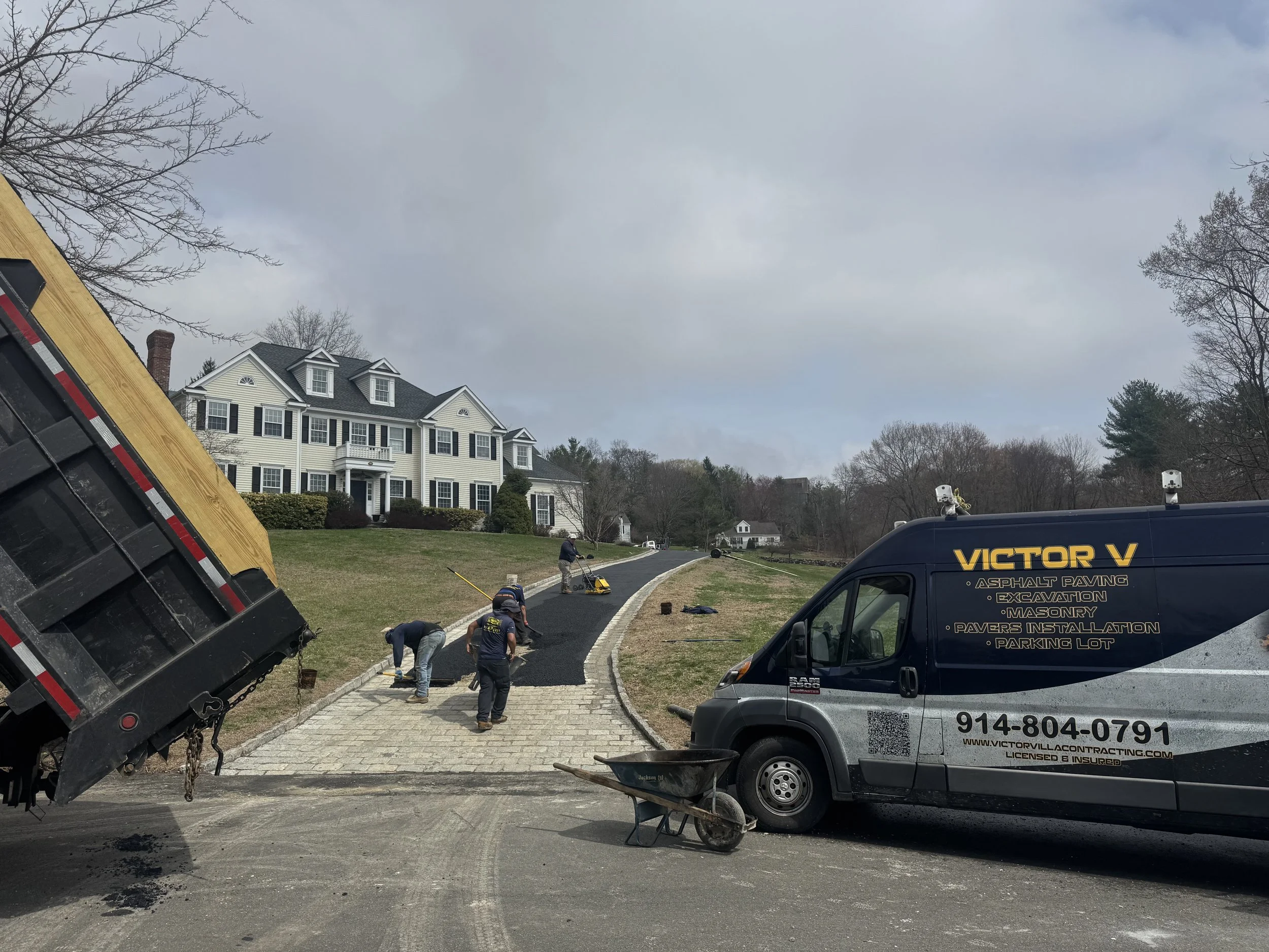 Workers paving a residential driveway with asphalt, with a large house in the background and a Victor V branded paving truck nearby.