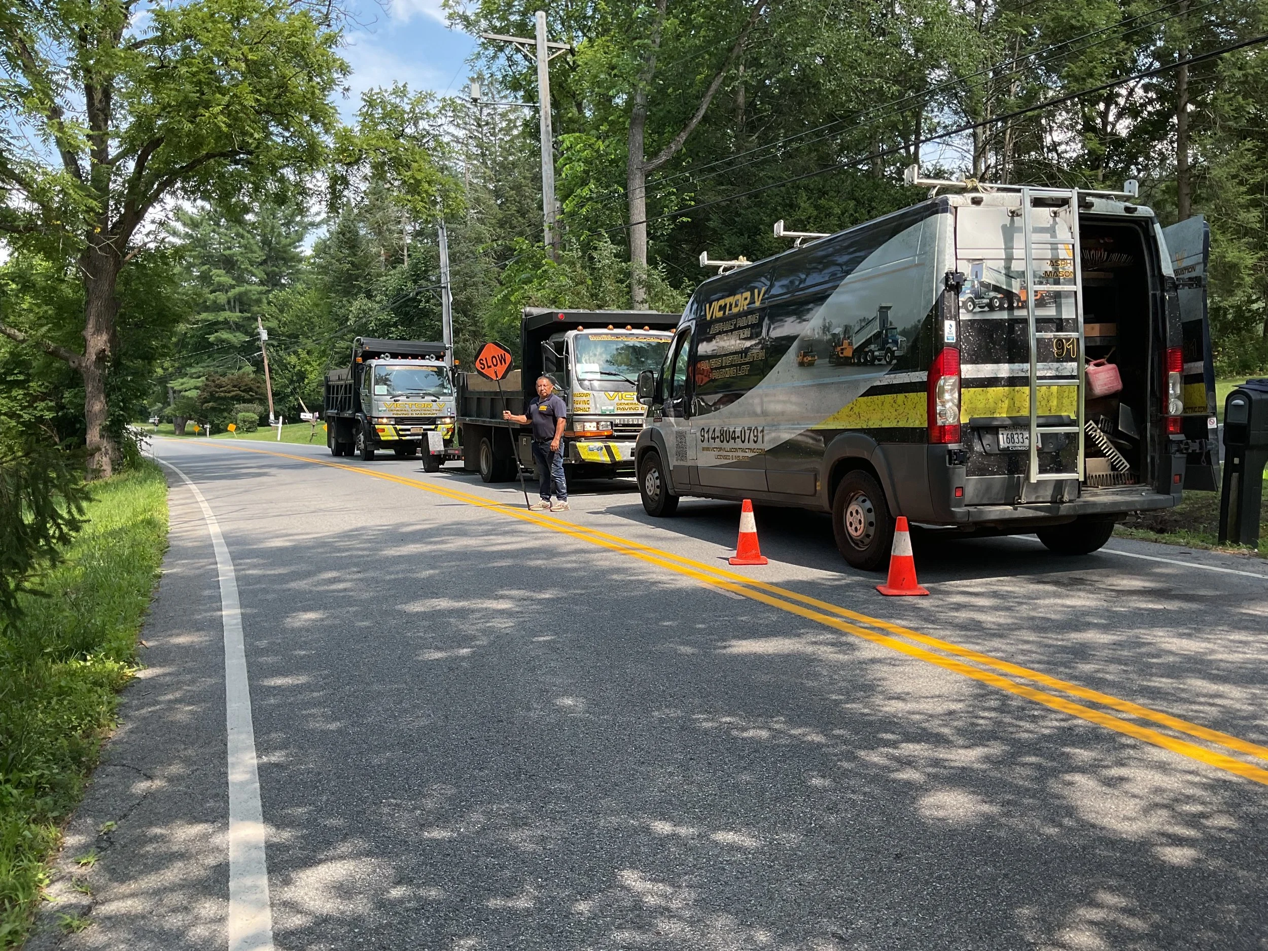 Line of construction or utility trucks parked on a rural two-lane road with orange cones, a worker standing by, and trees on both sides.