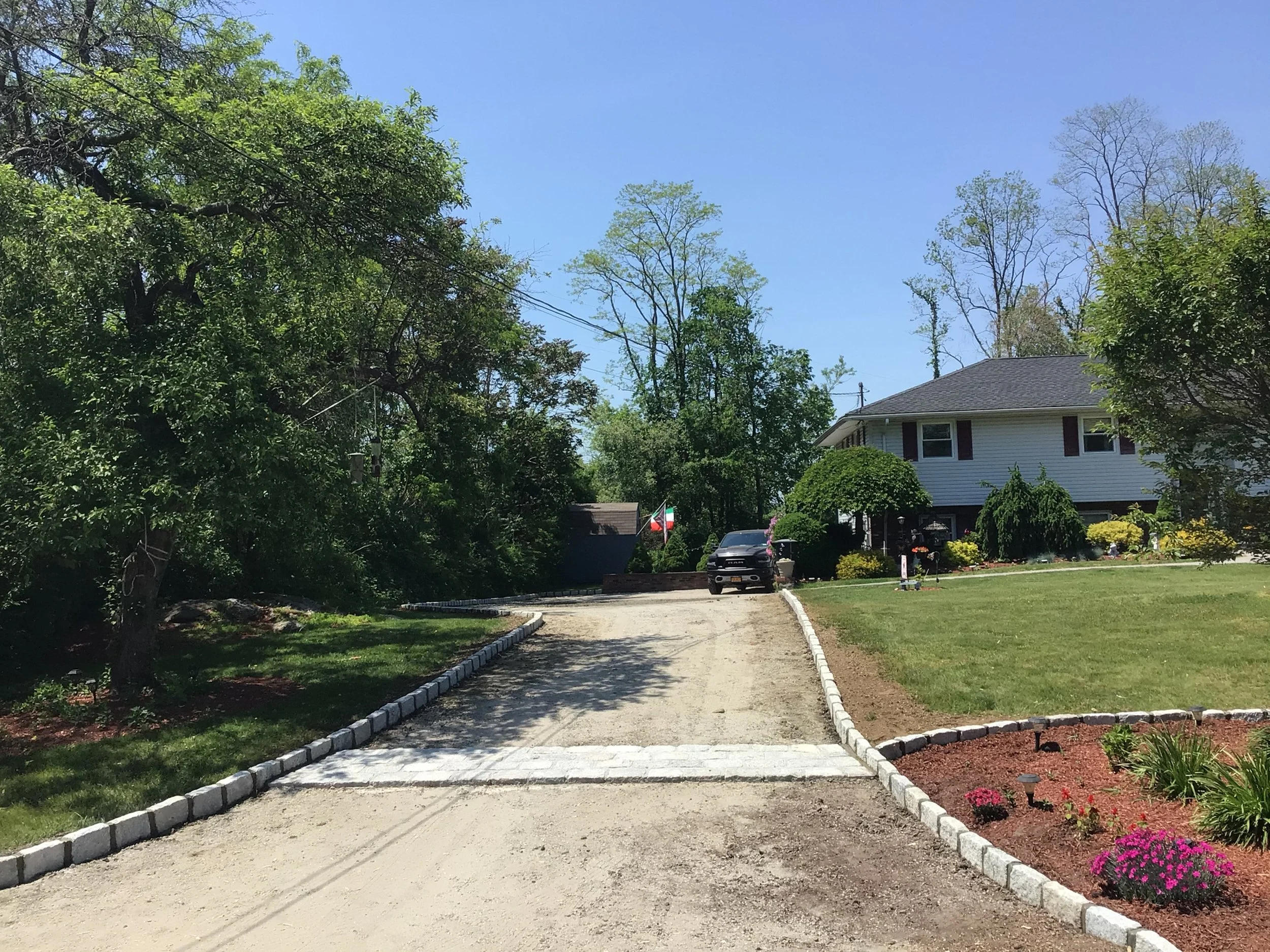 A dirt driveway leads to a house with a well-maintained garden and a parked car, surrounded by trees and a lawn under a clear blue sky.
