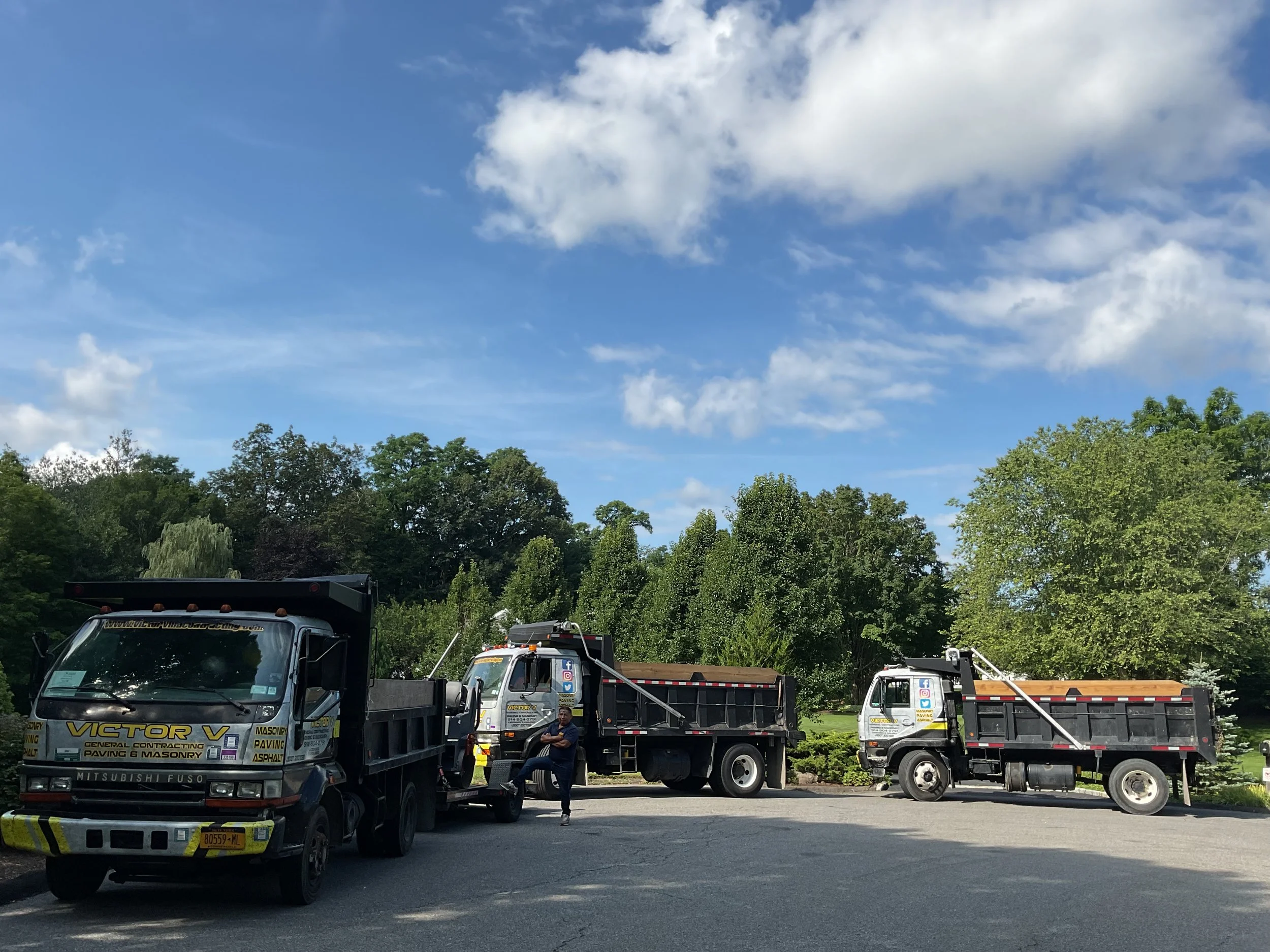 Three trucks parked in a lot with a person standing next to the middle truck, trees and a partly cloudy blue sky in the background.