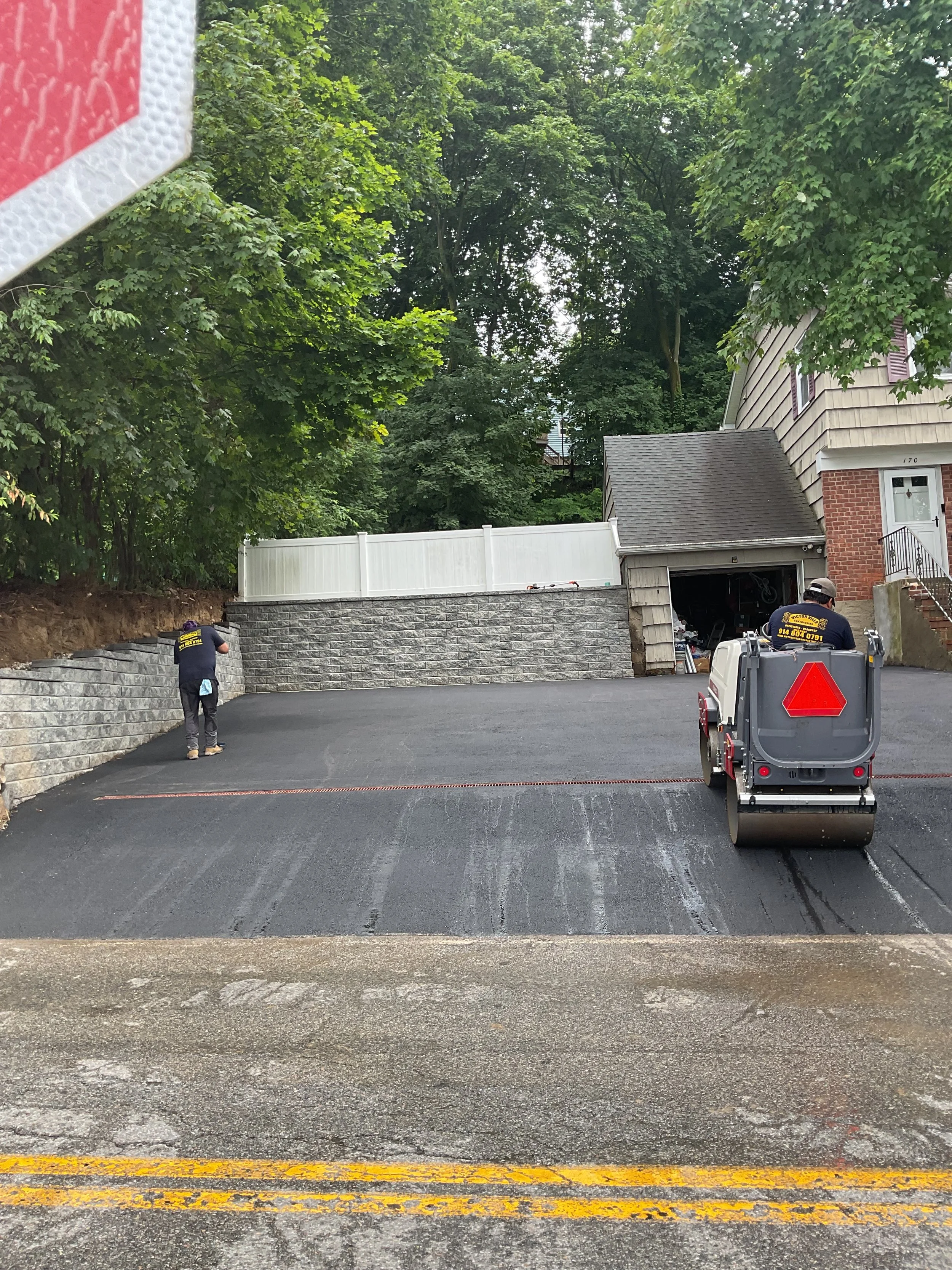 Workers resurfacing a driveway with asphalt, one operating a roller compactor and another using a tool near the left side, in front of a residential house with a garage.