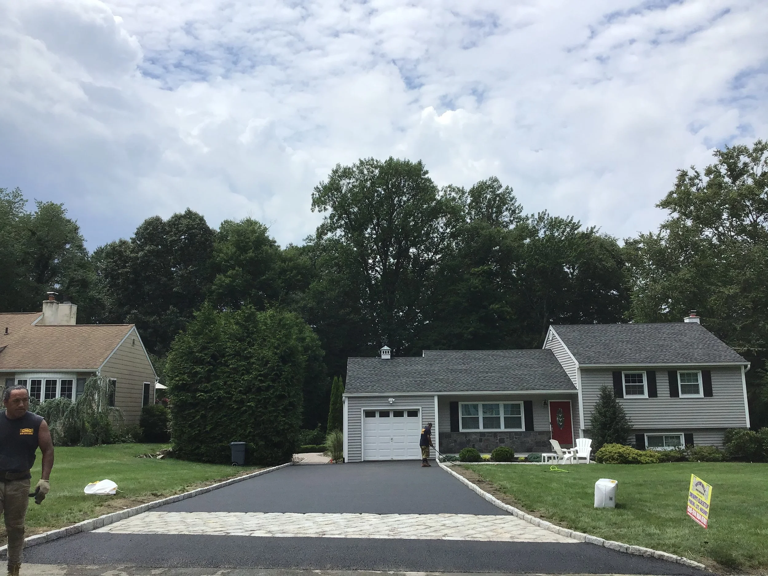 A house with a newly paved driveway in a suburban neighborhood, workers installing it, and a sign advertising asphalt sealing.
