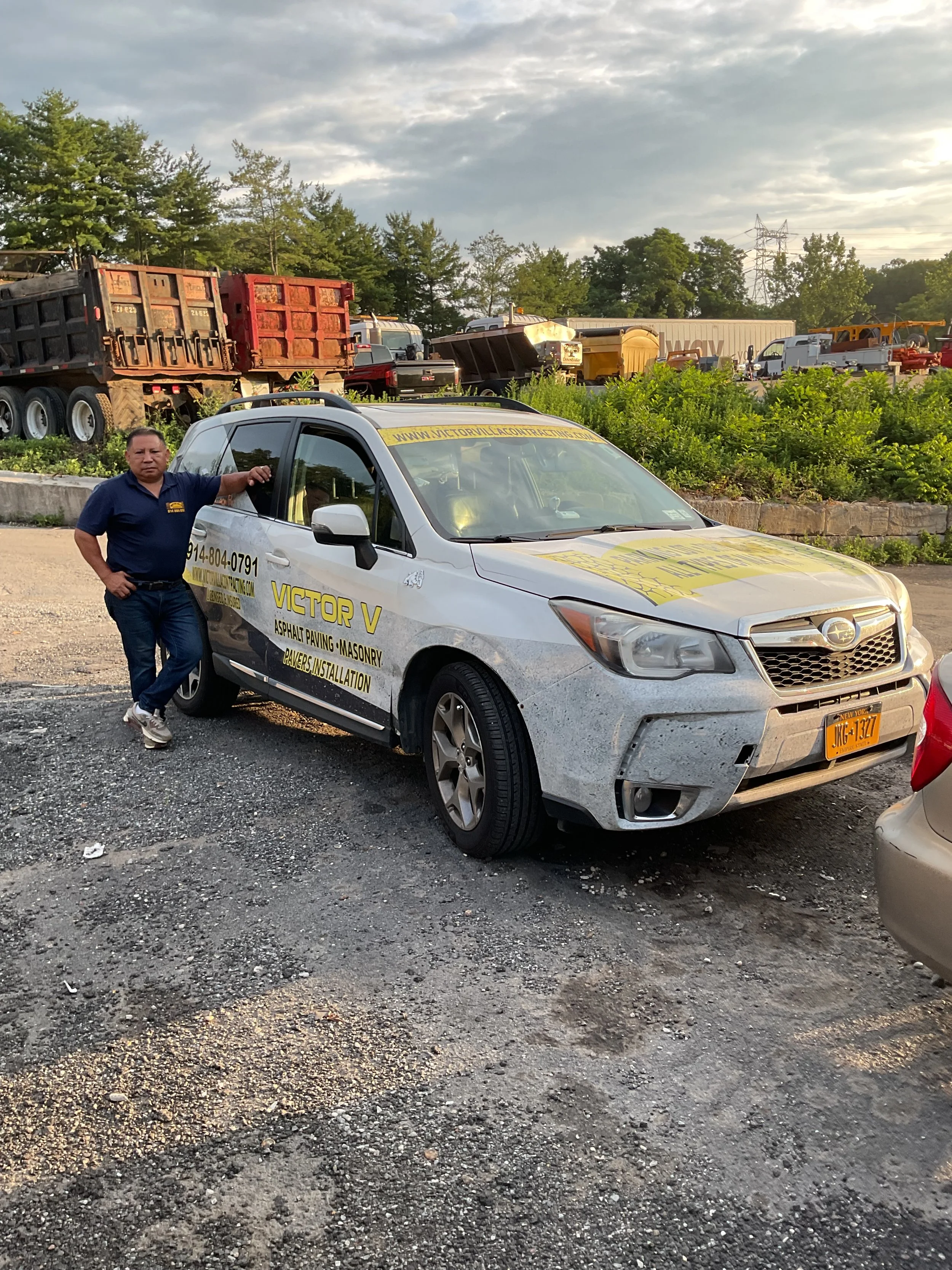 A man standing next to a white SUV with yellow signage, parked on a gravel lot, with construction equipment and vehicles in the background.