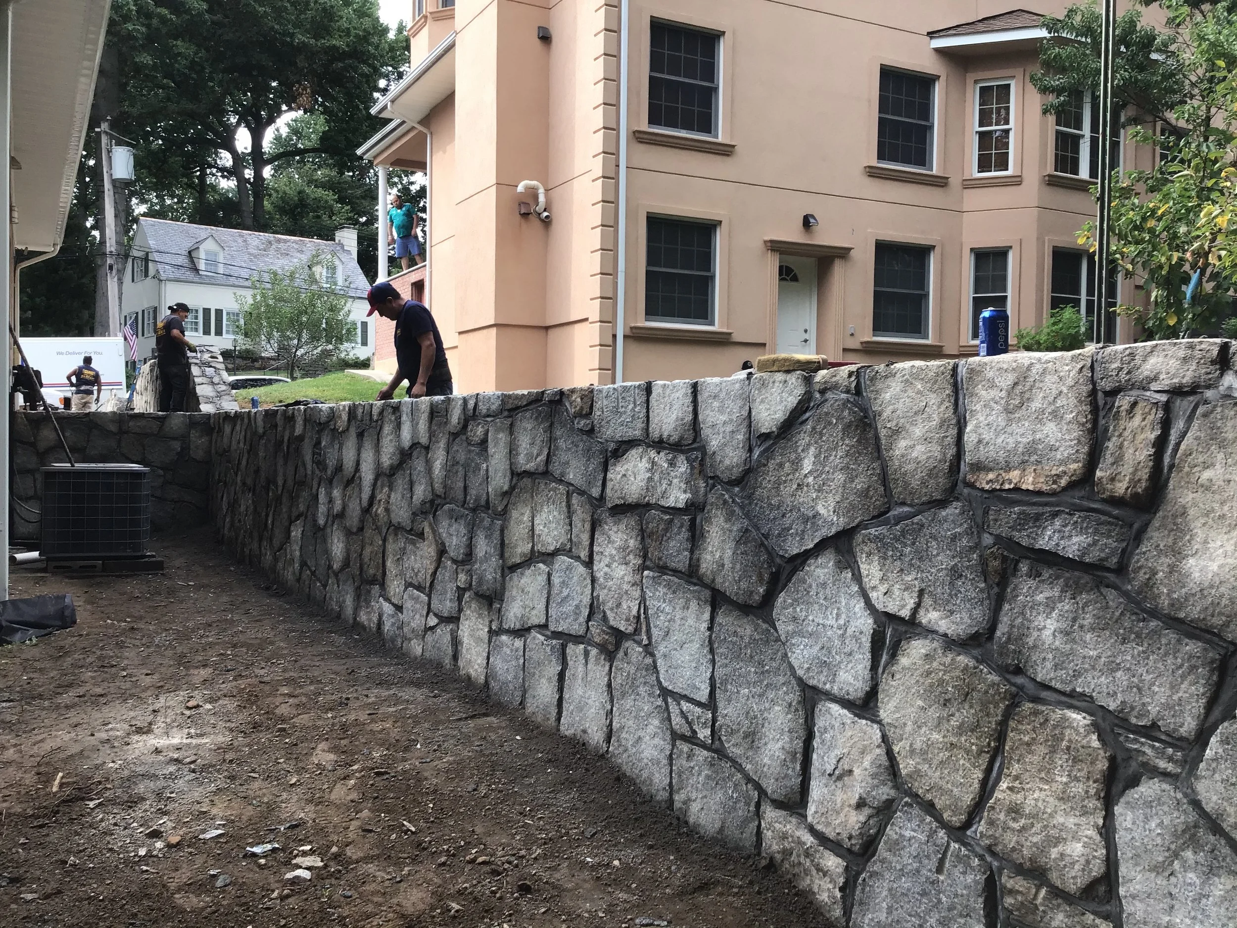 Workers constructing a stone retaining wall in a backyard with multi-story house and trees in the background.