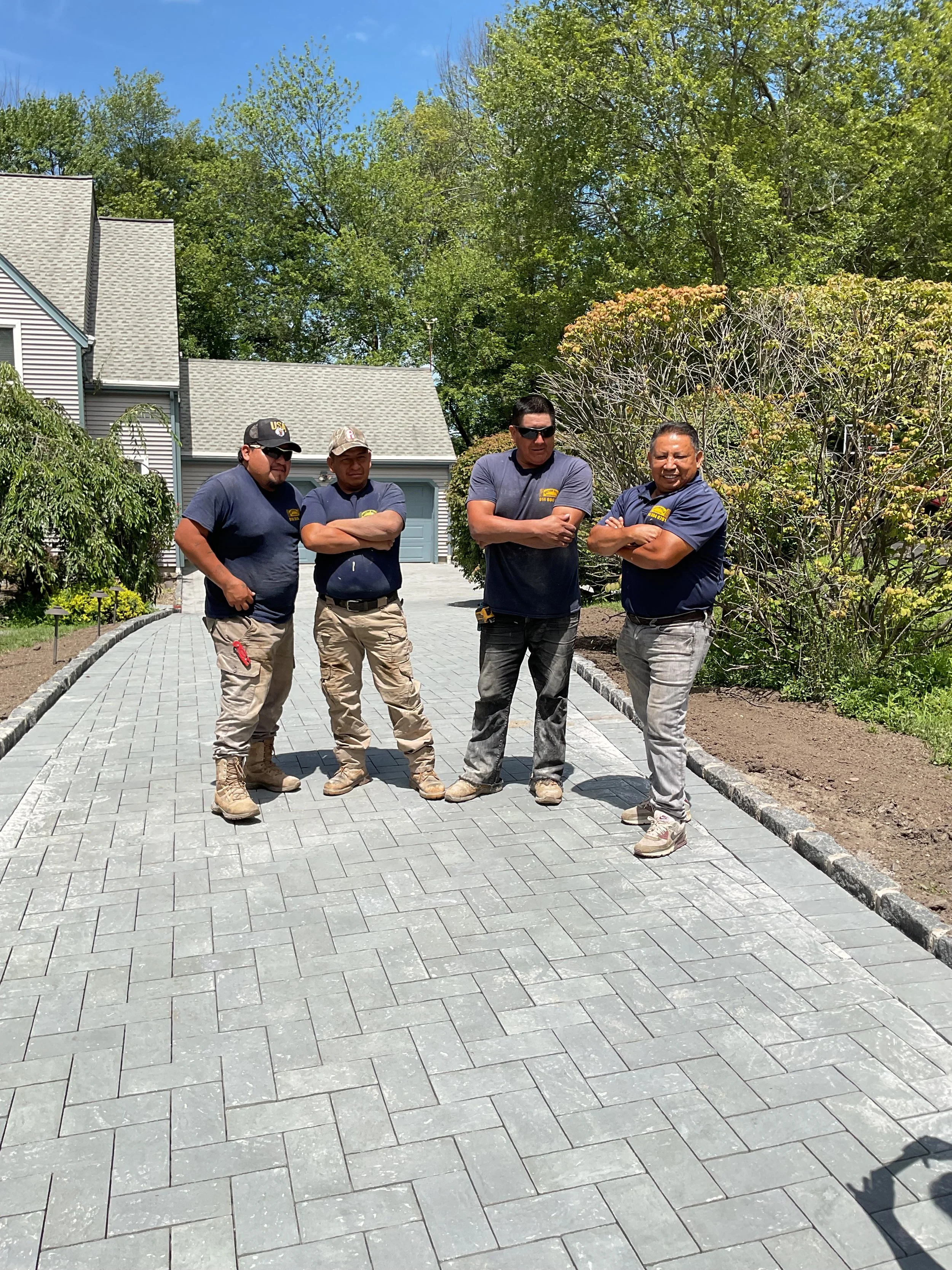 Four men standing on a newly paved driveway in front of suburban houses and green trees, arms crossed, wearing work clothes.