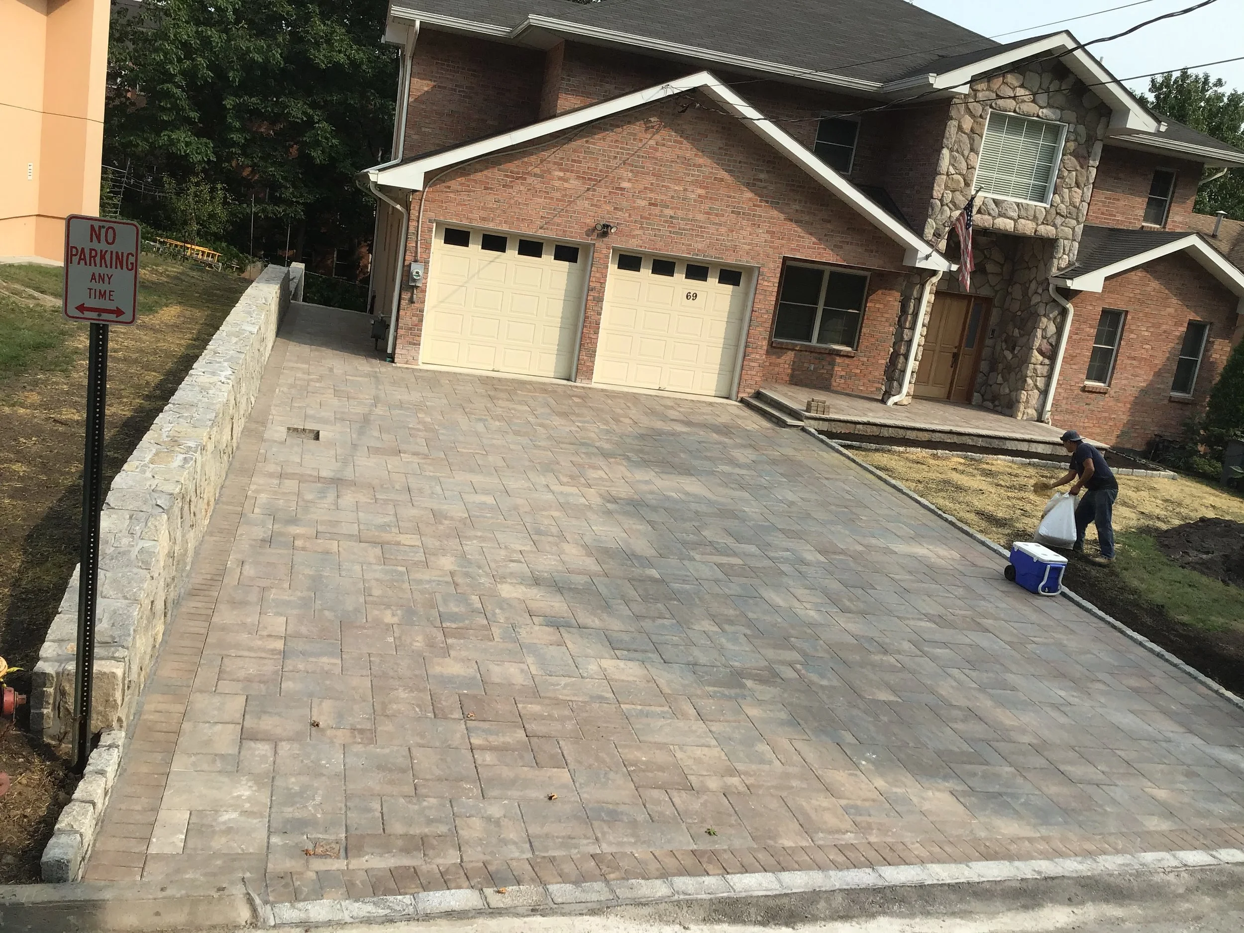 A worker laying down bricks on a newly paved driveway in front of a brick house with a stone facade over the entrance. There is a stone retaining wall on the left side with a no parking sign, and the worker is placing bricks along the edge of the pav