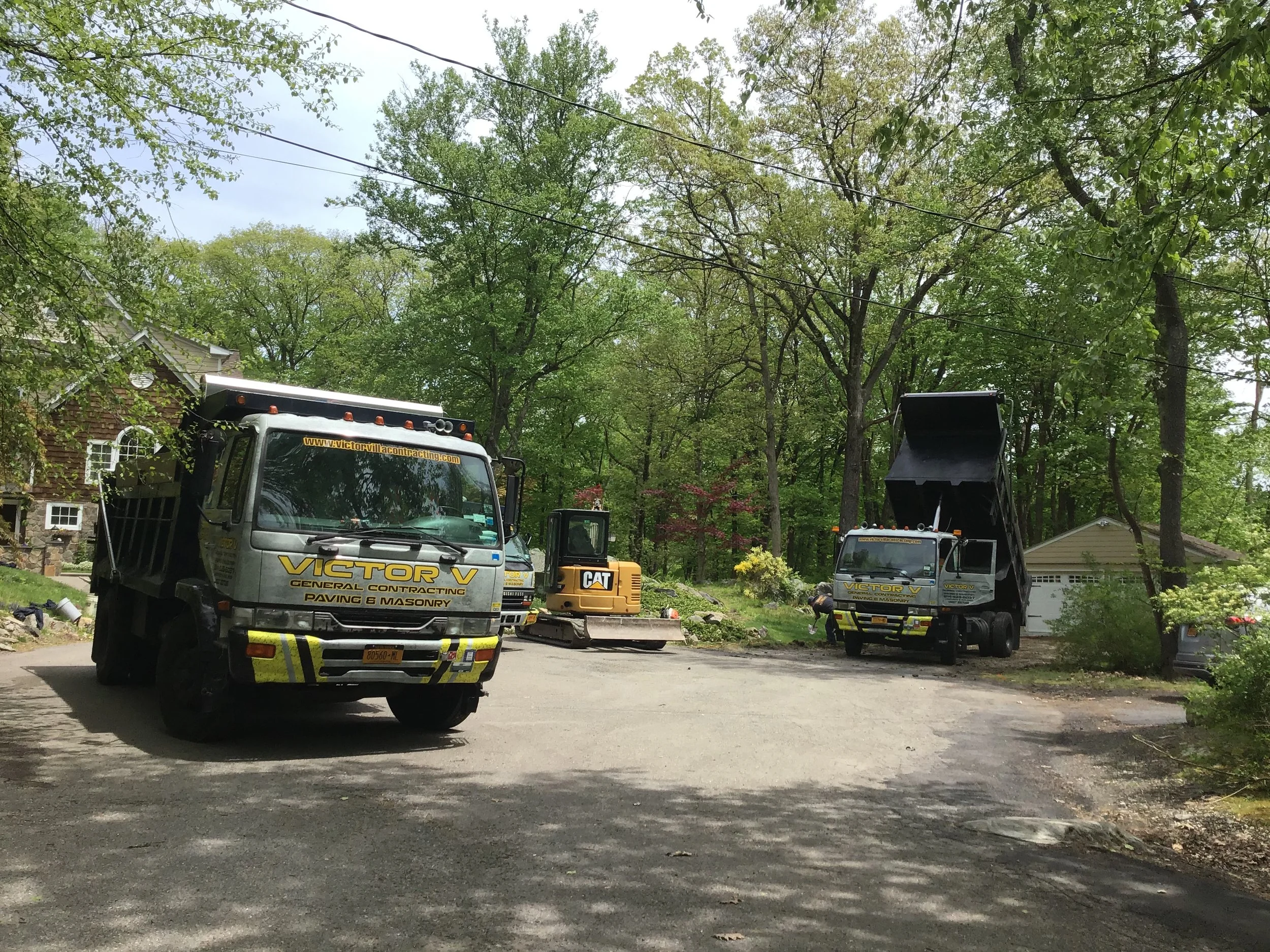 Construction workers and trucks working on a residential road surrounded by trees, with one truck having its bed raised.