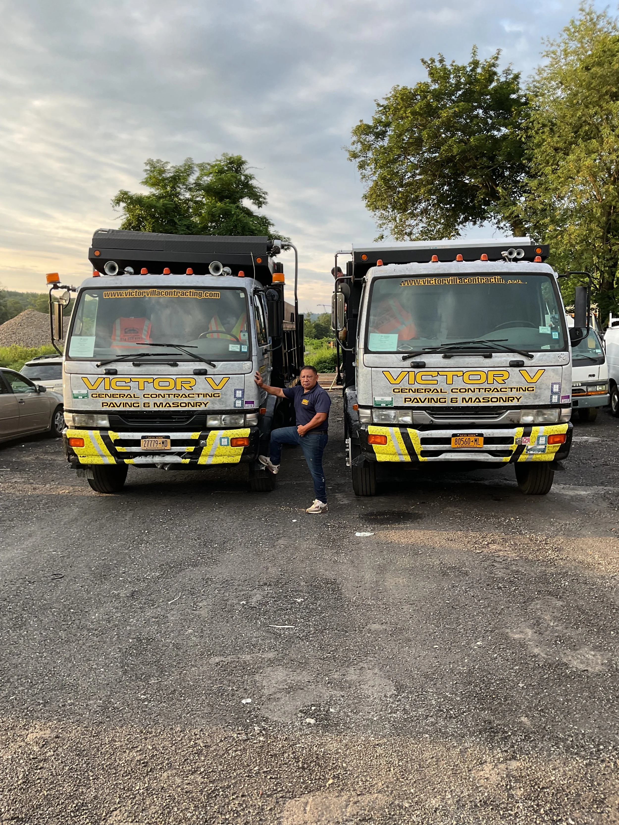 Two construction trucks labeled 'Victor V General Contracting Paving & Masonry' parked side by side on a gravel lot. A man is leaning against the front of the left truck, pointing towards the camera. Trees and a partly cloudy sky are in the backgroun