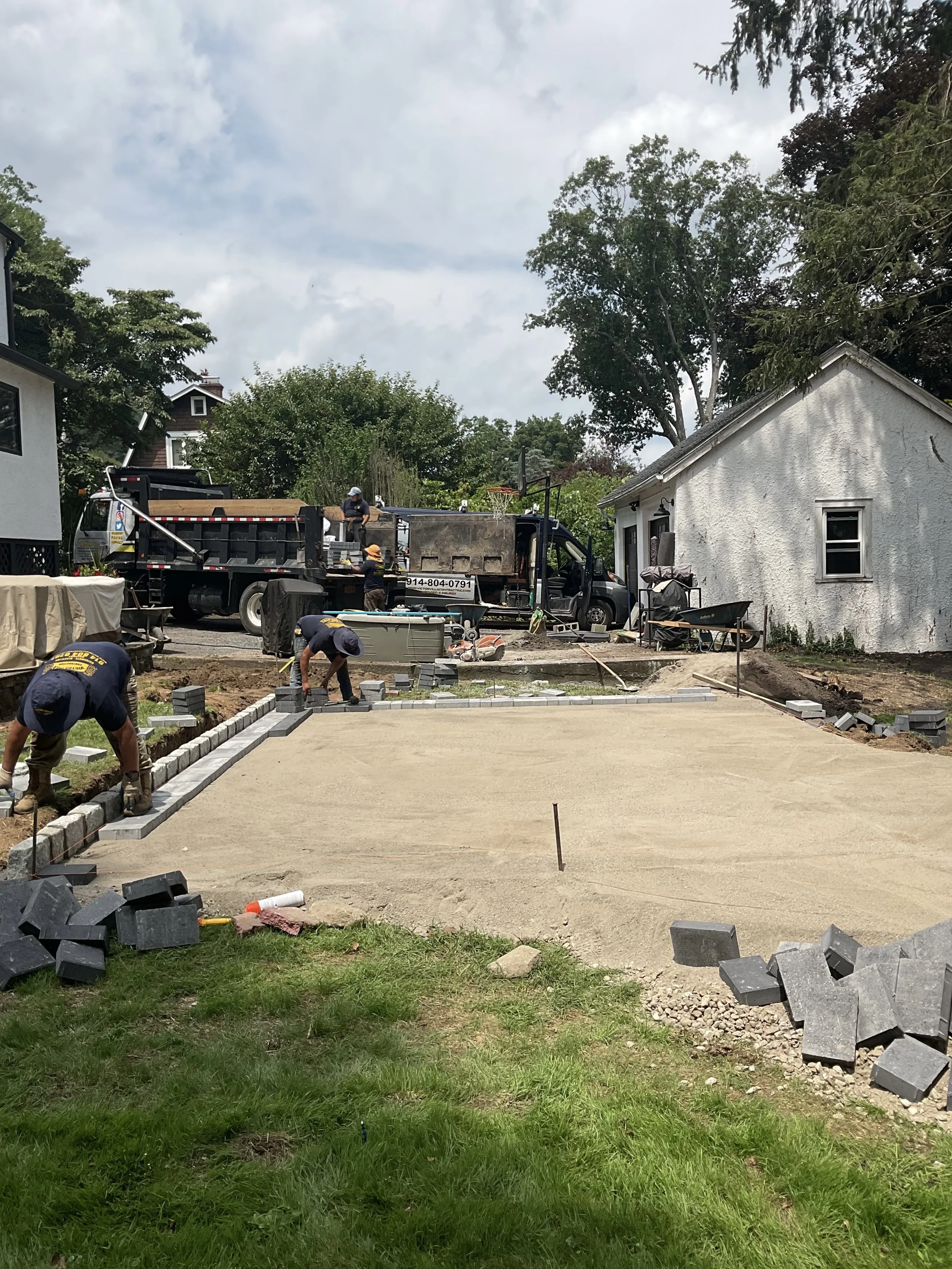 Construction workers laying pavers to create a walkway or patio in a residential backyard, with a truck and a house nearby.