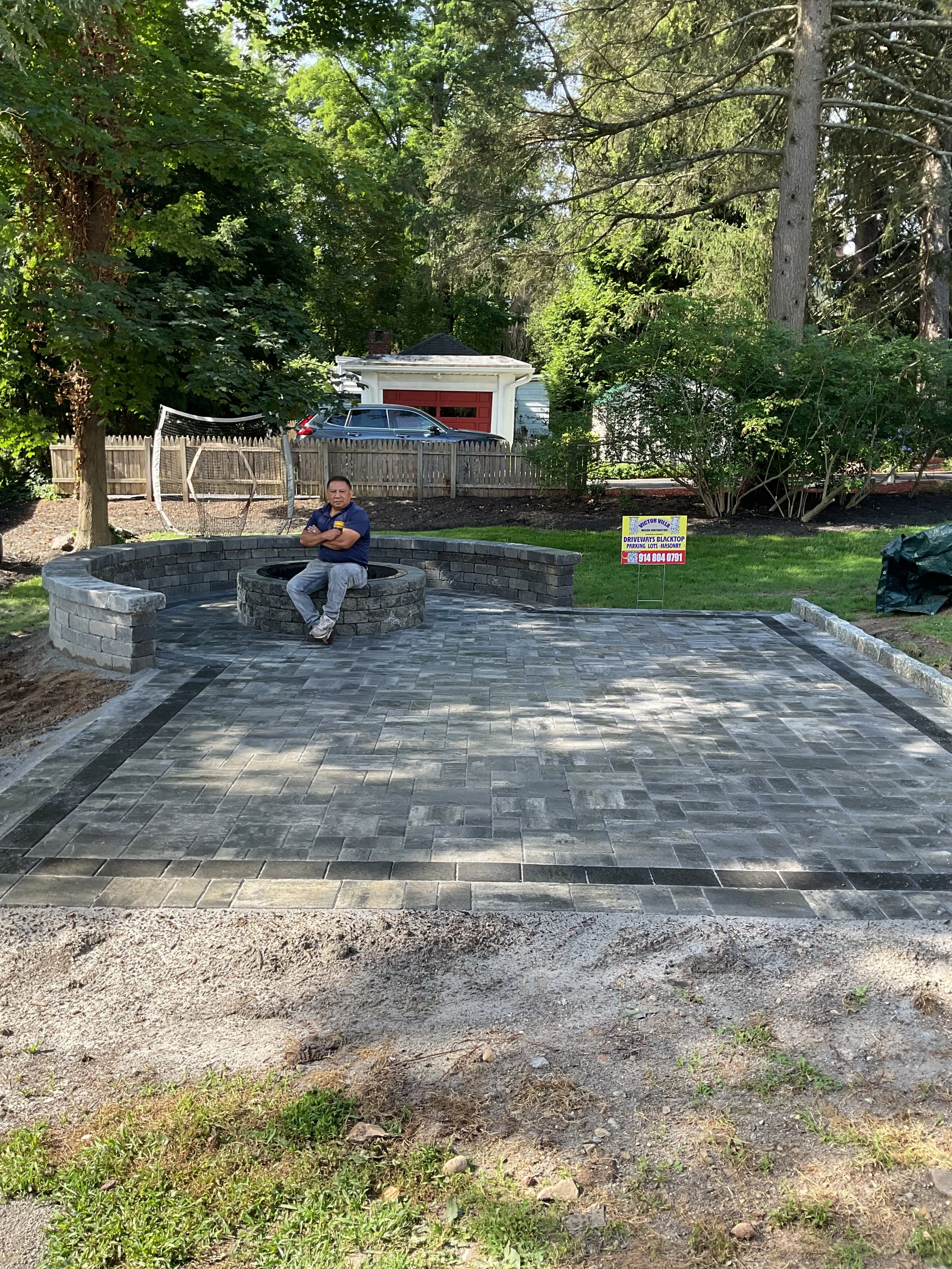 Man sitting on a circular brick bench in a backyard with newly paved stone patio, surrounded by trees and a wooden fence.