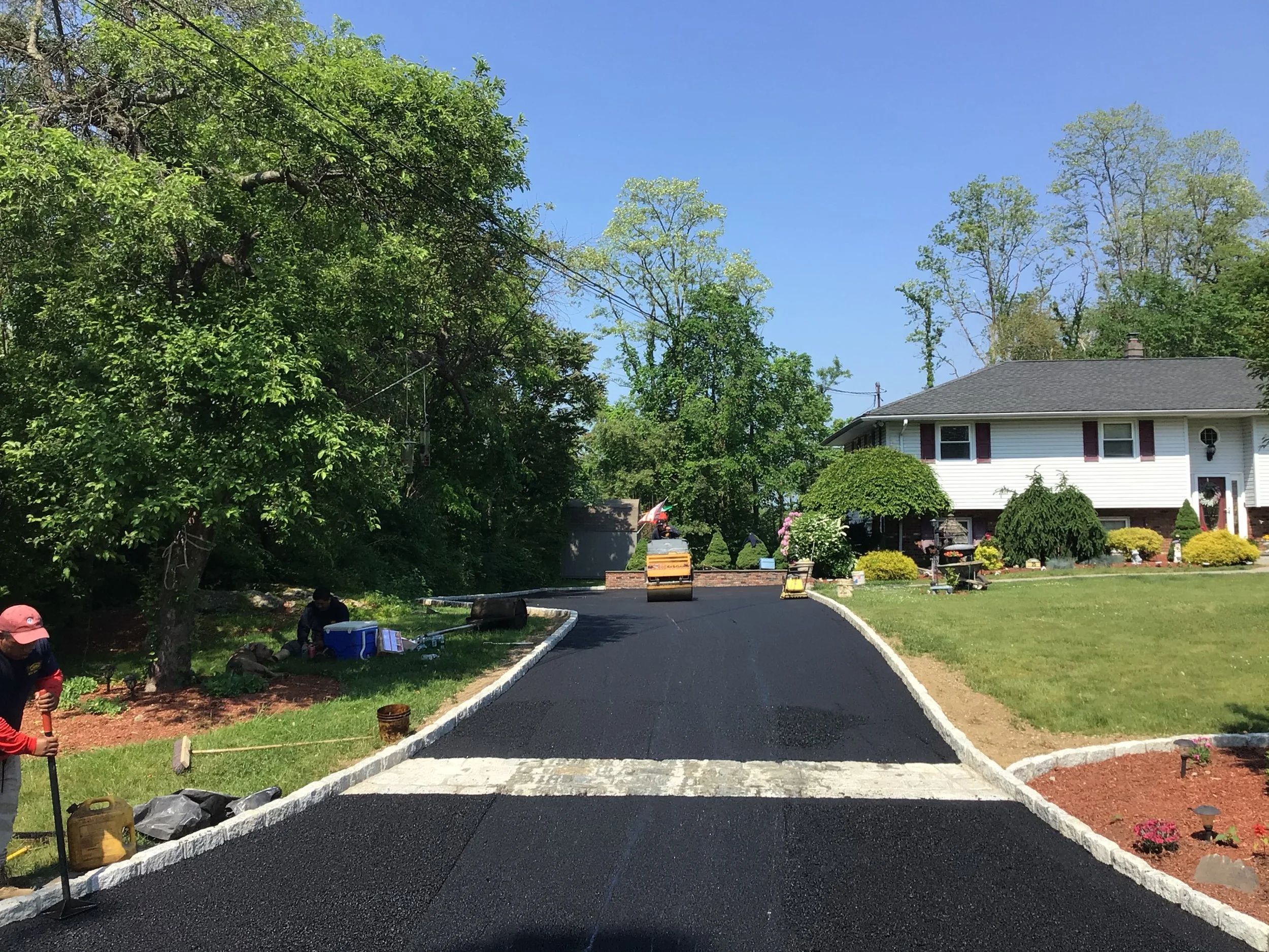 A newly paved driveway leading to a white house with a gray roof and a landscaped front yard. Two workers are working on the side of the driveway under trees, with construction equipment and materials visible.