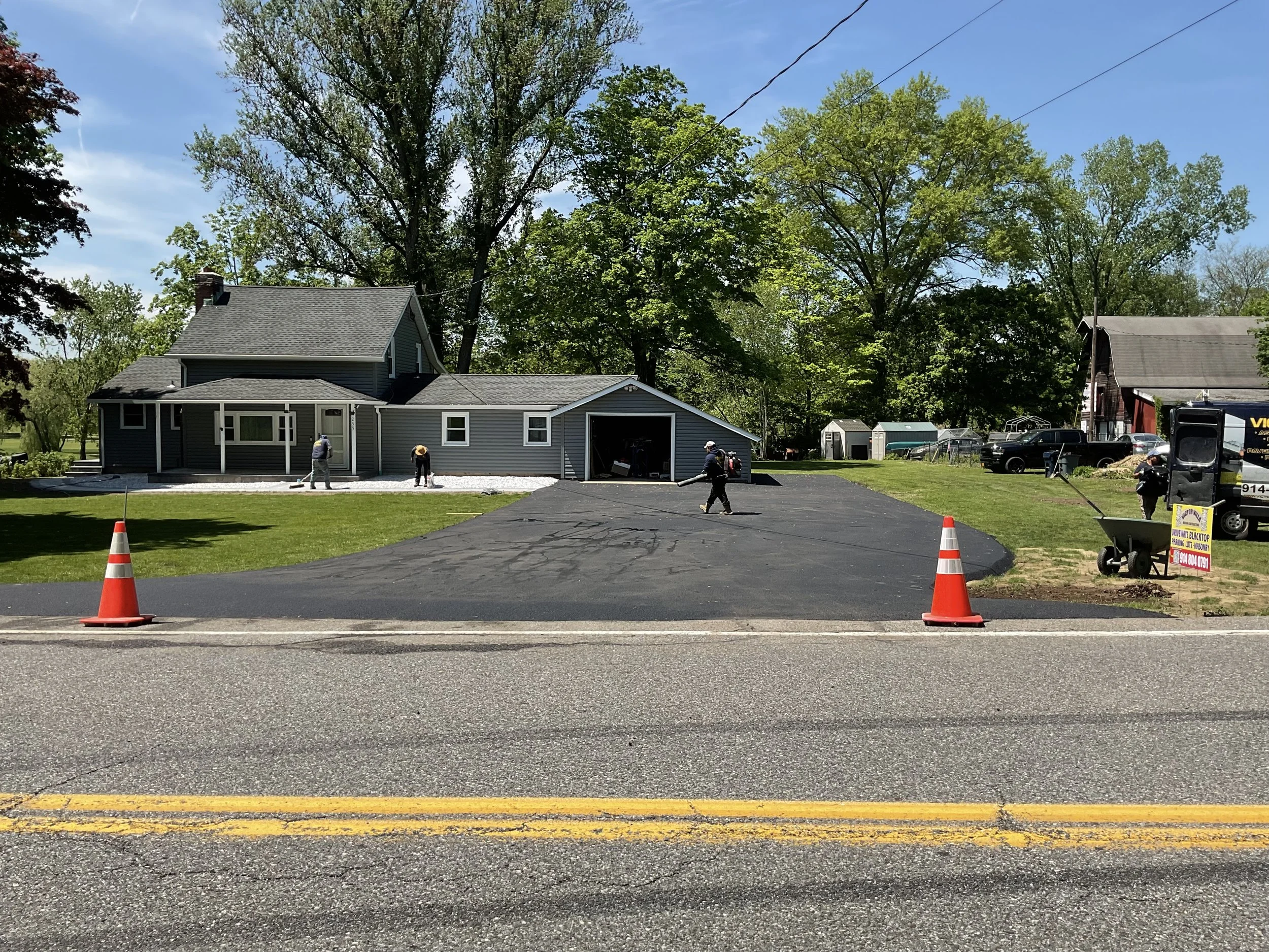 Homeowner is paving a driveway with black asphalt, with construction workers working on it, parking cones indicating construction, next to a house with trees in the background.