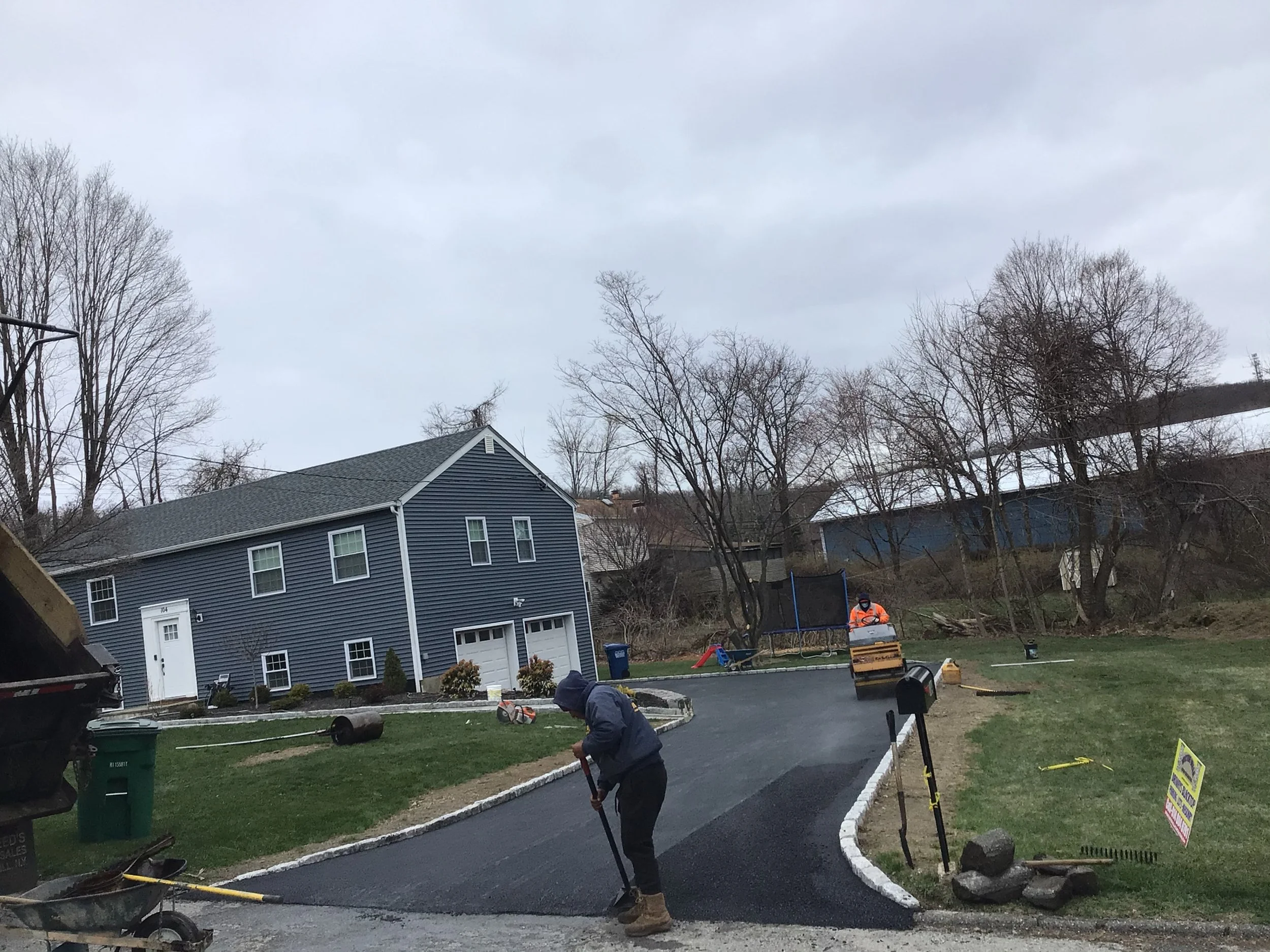 Workers paving a driveway in front of a blue house on an overcast day.