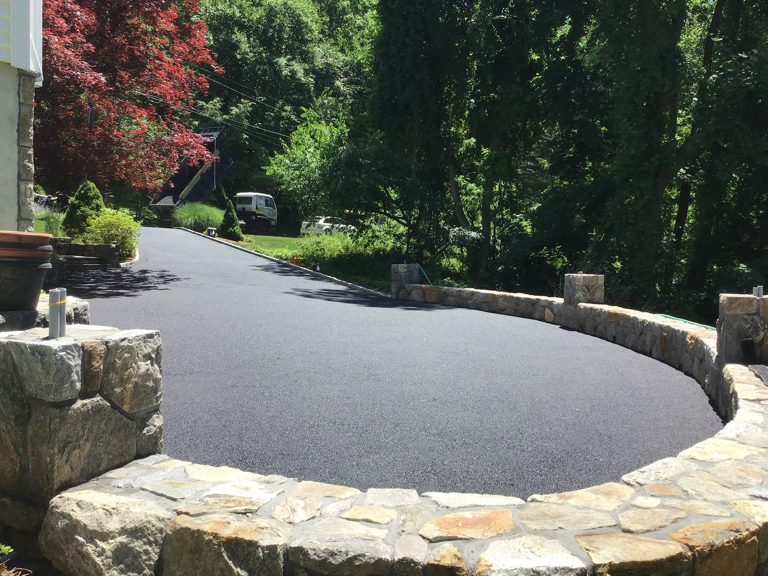 Freshly paved asphalt driveway with a stone border, surrounded by green trees and shrubs, with a red-leafed tree on the left and a white truck in the background.