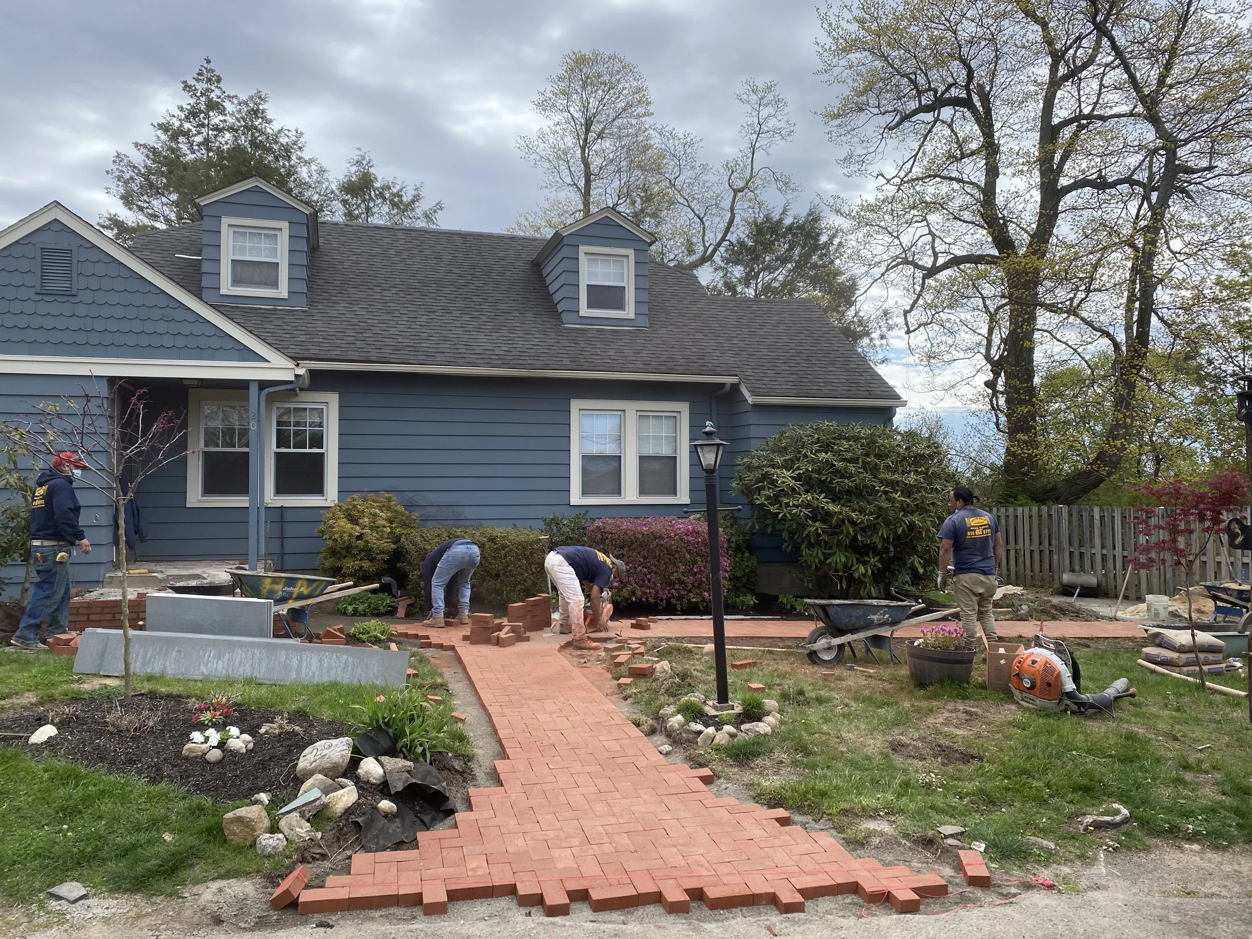 Construction workers laying red brick pavers for a walkway in the front yard of a blue house with dormer windows, surrounded by trees and garden plants.