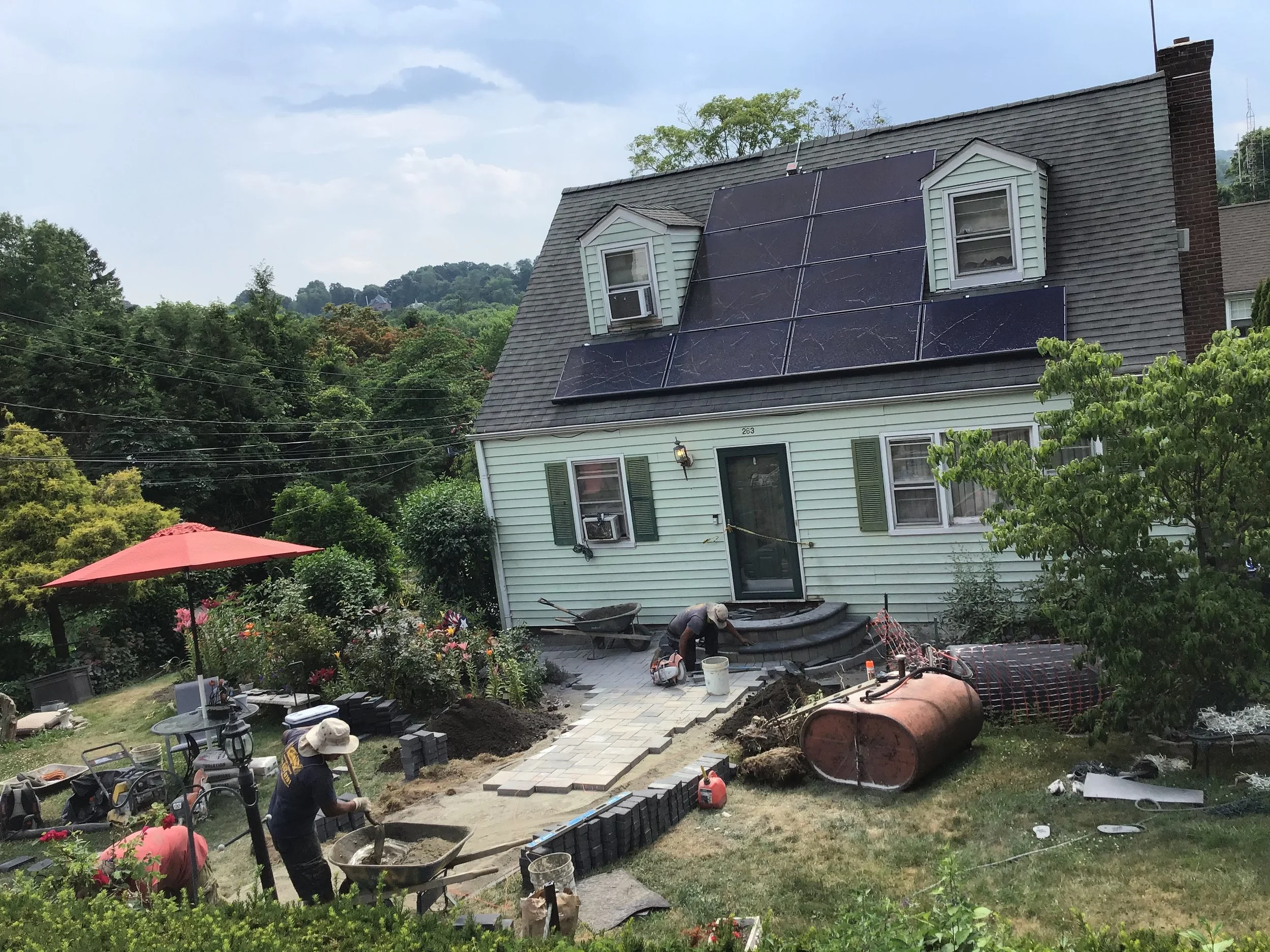 A house with solar panels being installed on the roof. Workers are laying bricks and working on the patio area in the backyard, which includes flowers, a red umbrella, and construction equipment.