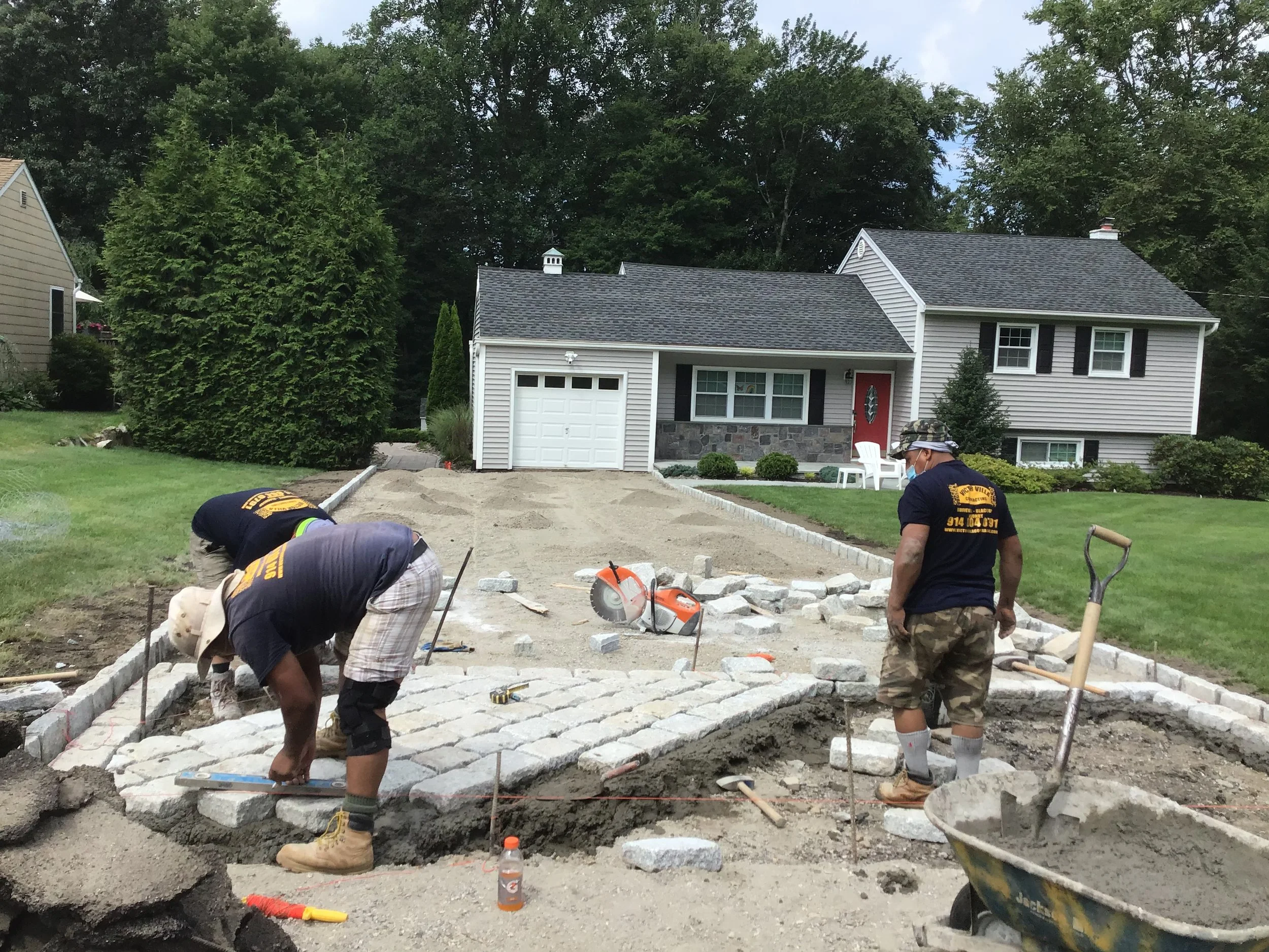 Three workers laying stones in a driveway in front of a suburban house with a driveway and lawn.