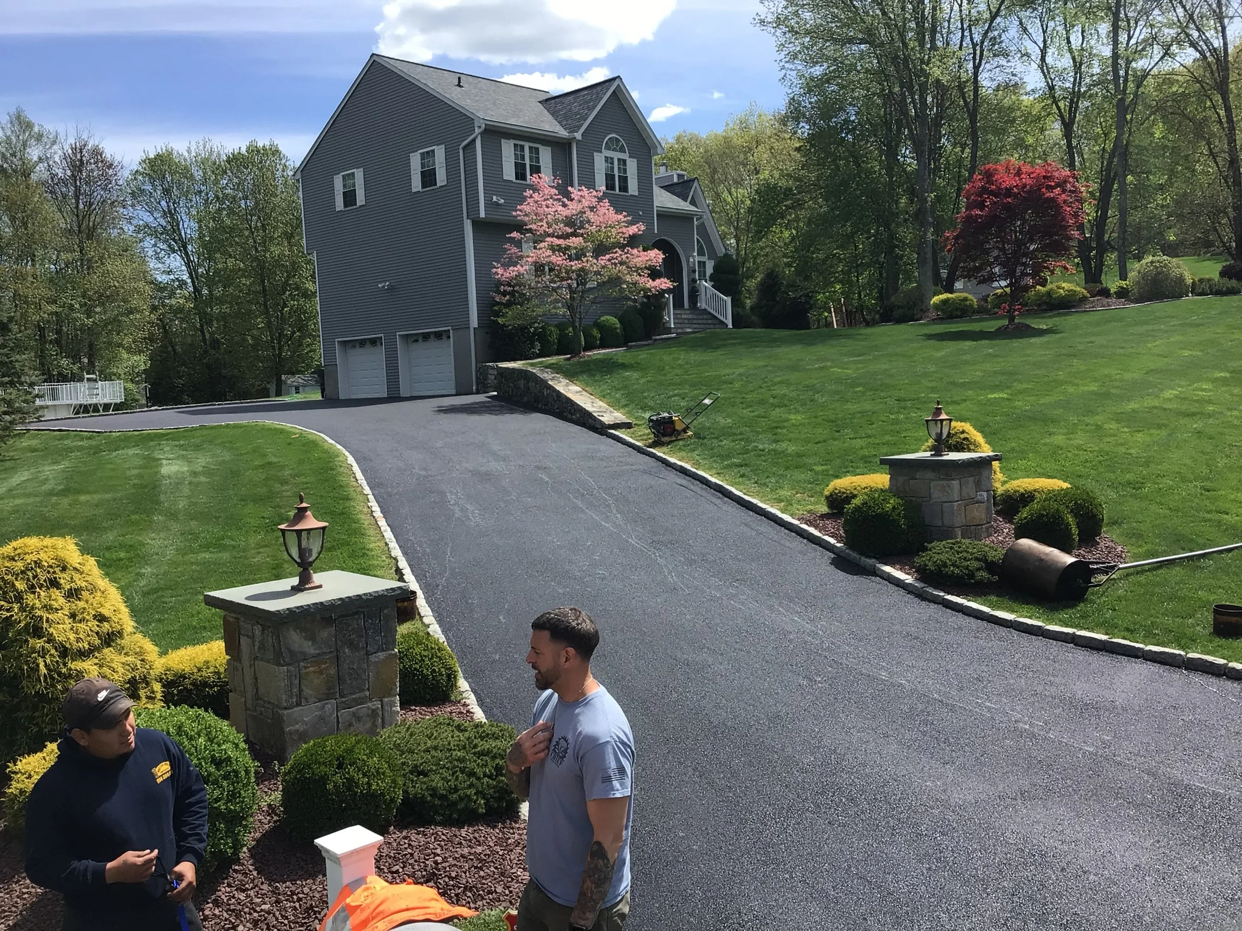 Two men standing near a newly paved driveway in front of a large house with well-maintained lawn and landscaping, including flowering trees and shrubs.